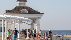 A pier with a gazebo structure, people walking, and a beach in the background.