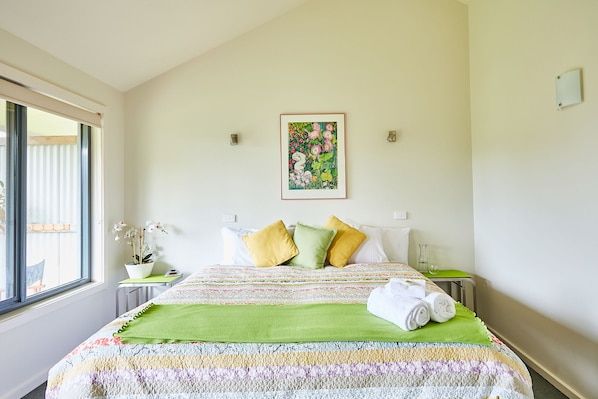 A bedroom with a large bed, a window, a potted plant, and a framed floral artwork on the wall.