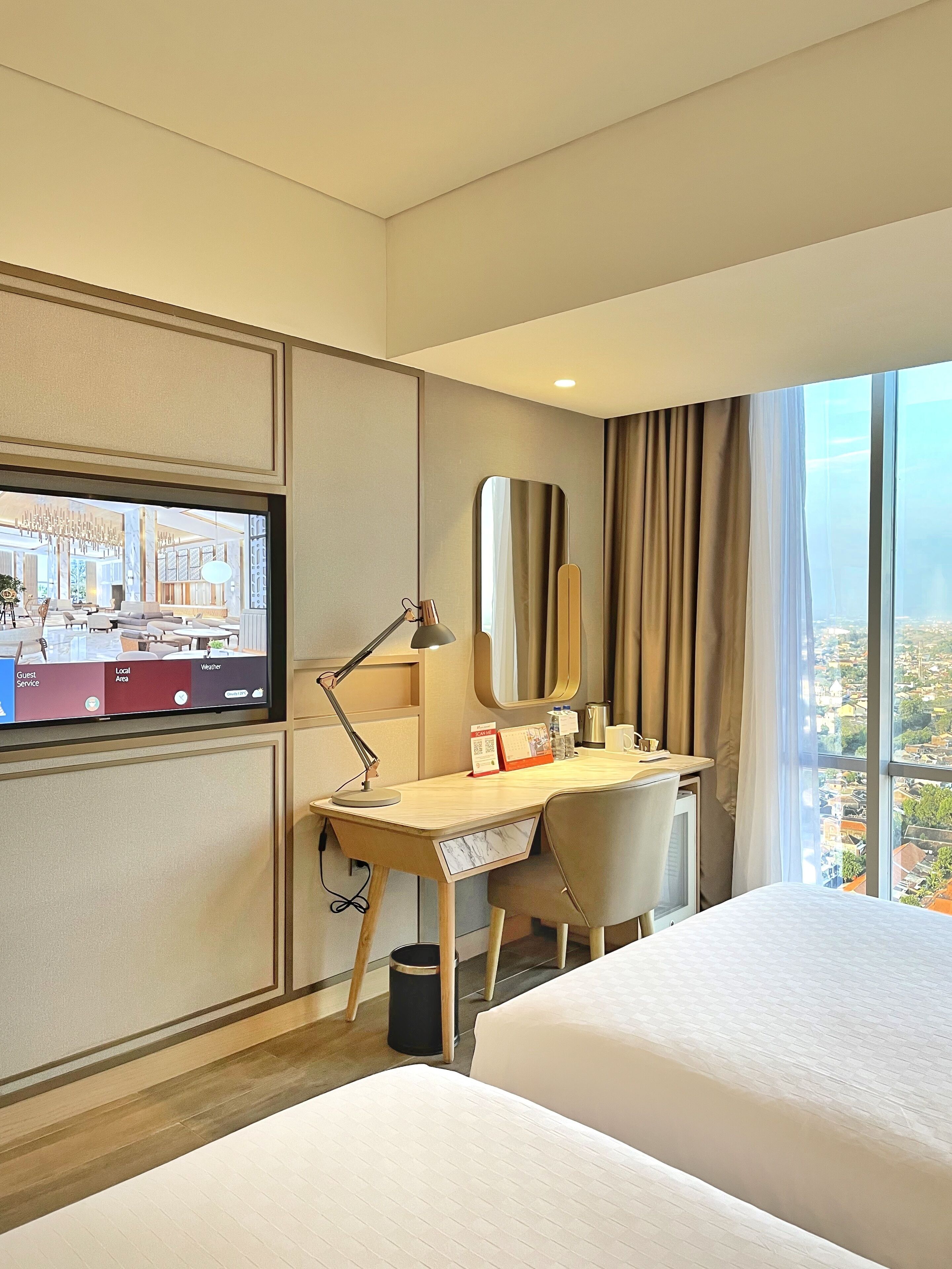 A hotel room with a bed, desk, chair, TV, and a large window with a city view.