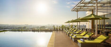 A rooftop infinity pool with sun loungers and umbrellas, overlooking a cityscape.