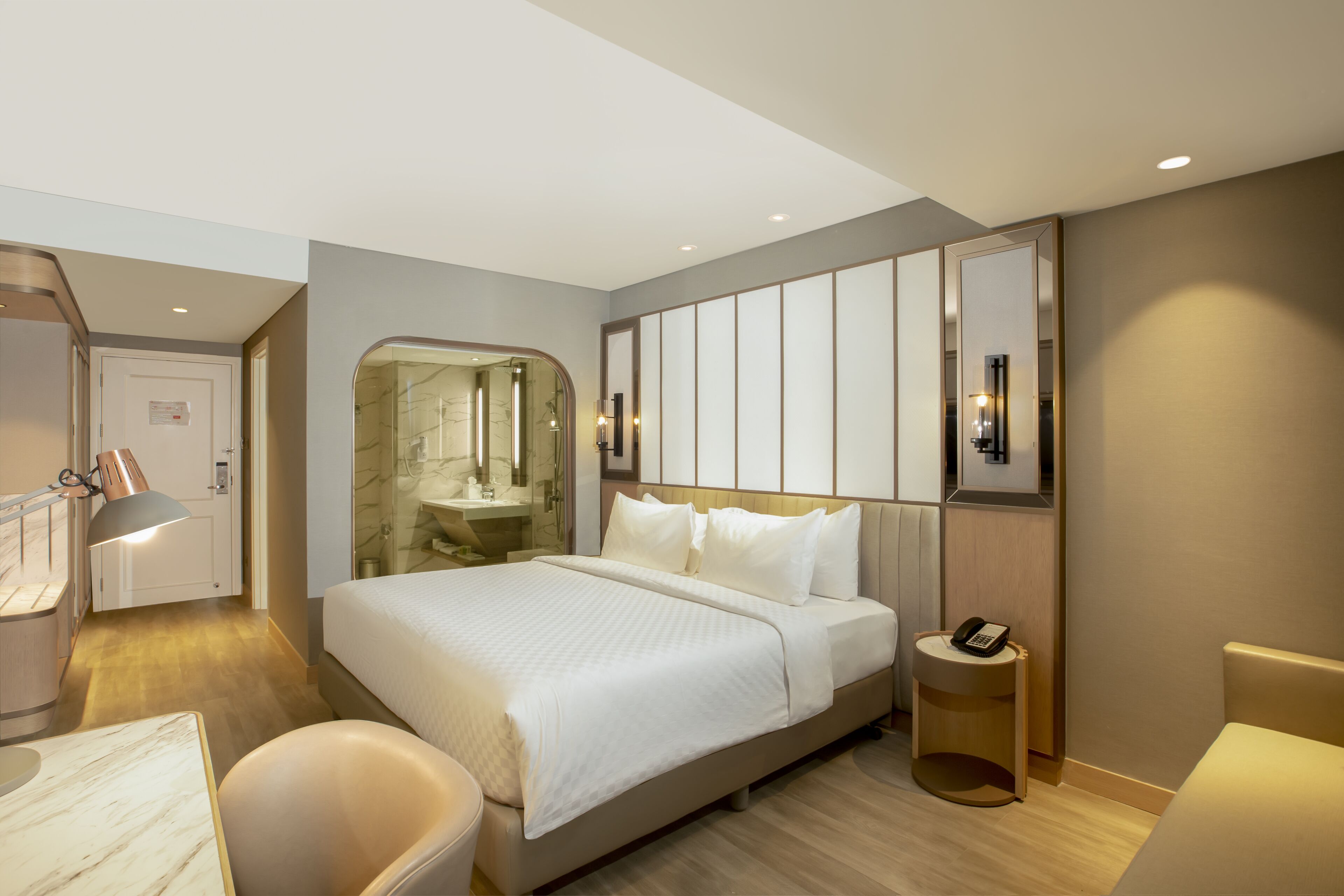 A modern hotel room with a large bed, a bathroom with a glass shower, a bedside table with a phone, and a seating area with a chair.