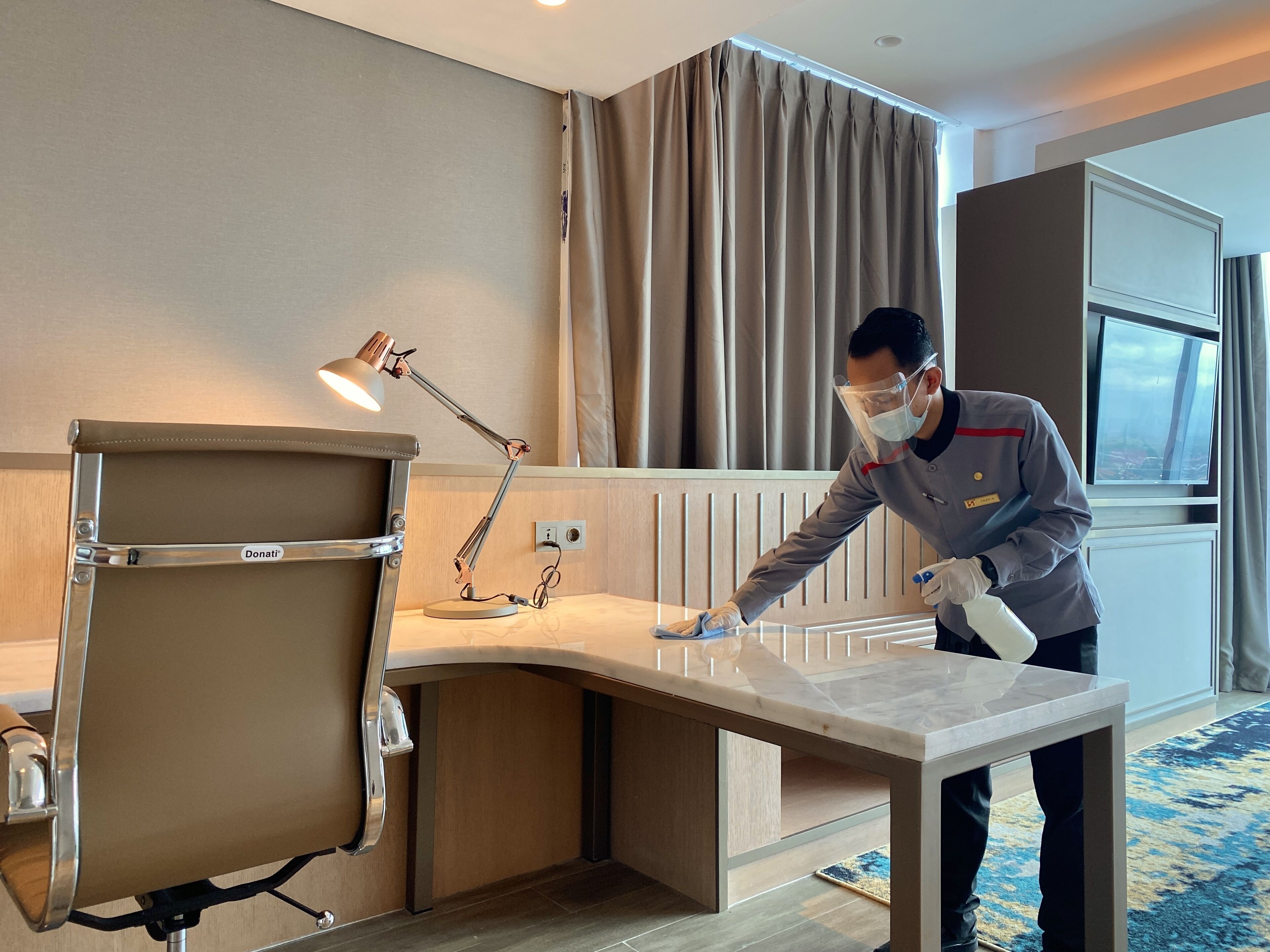 Een hotelkamer met een bureau, een lamp en een persoon die het bureau schoonmaakt.
