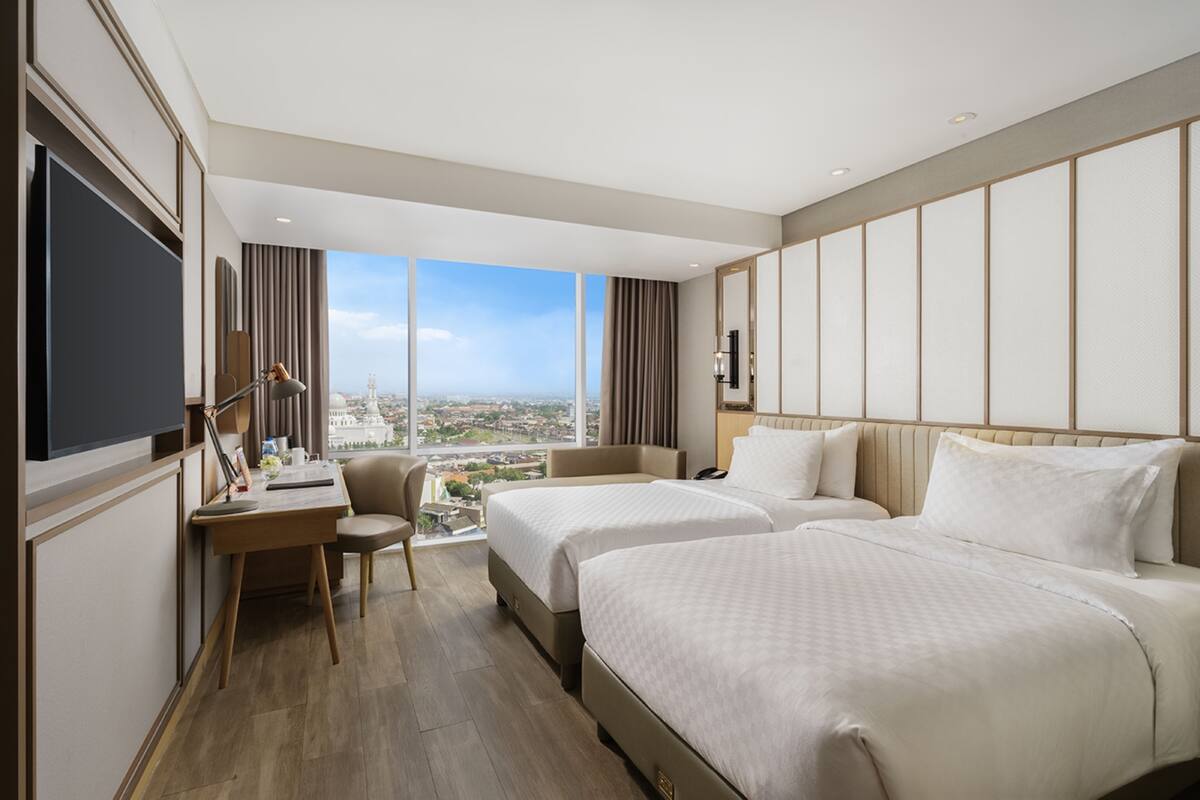 A hotel room with two beds, a desk, and a chair. There is a large window offering a city view.