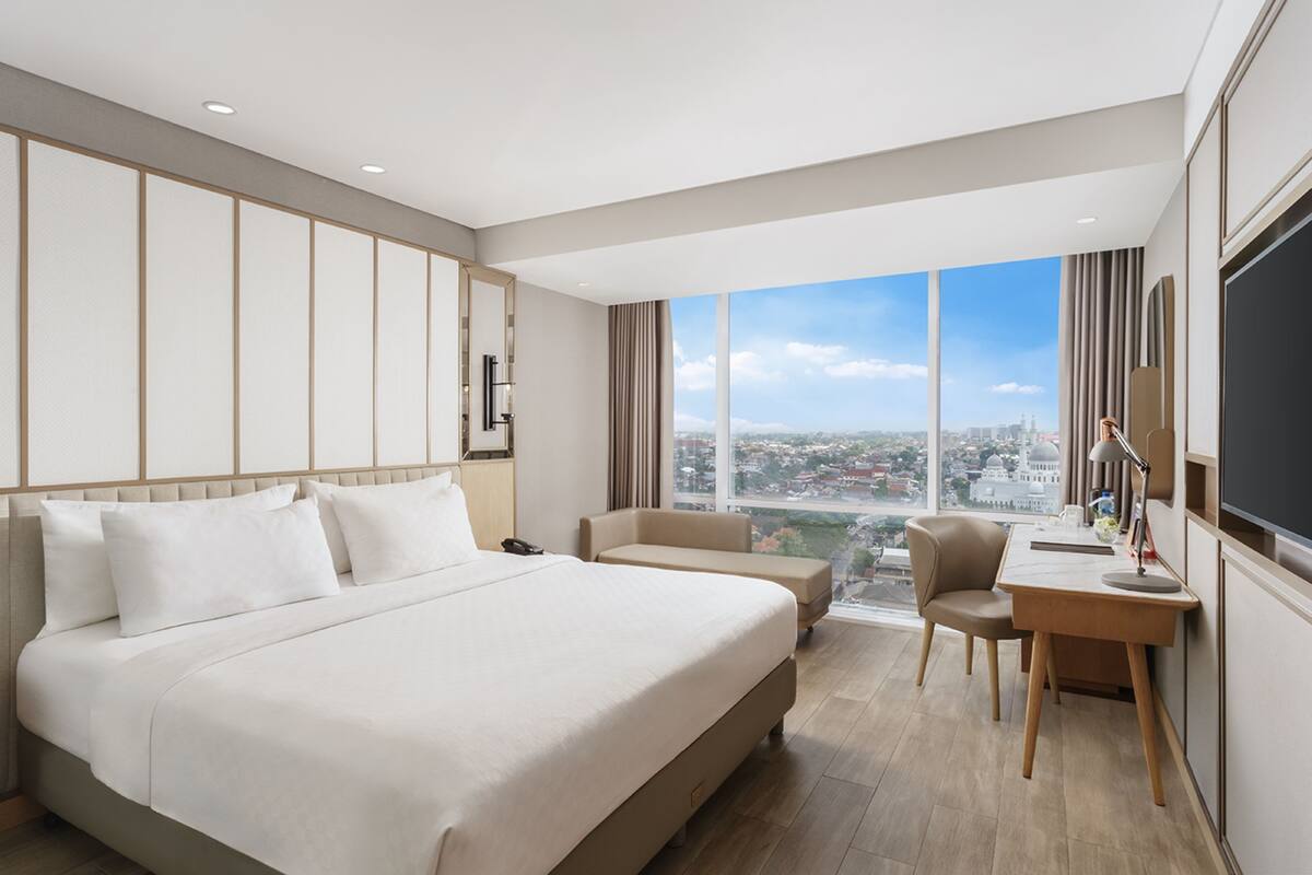 A modern hotel room with a large bed, a desk, and a view of the cityscape.