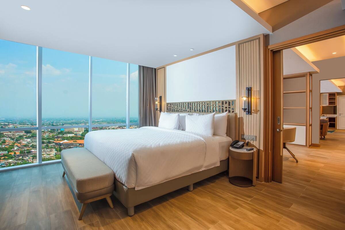 A modern hotel room with a large bed, a bench, a bedside table with a phone, and a view of the city.