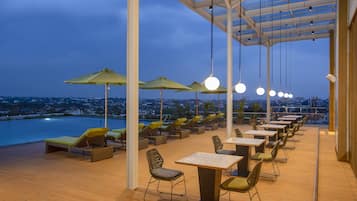 Zona de lounge en la azotea con sillones de sol, mesas y sombrillas, con vistas a un paisaje urbano y una masa de agua.