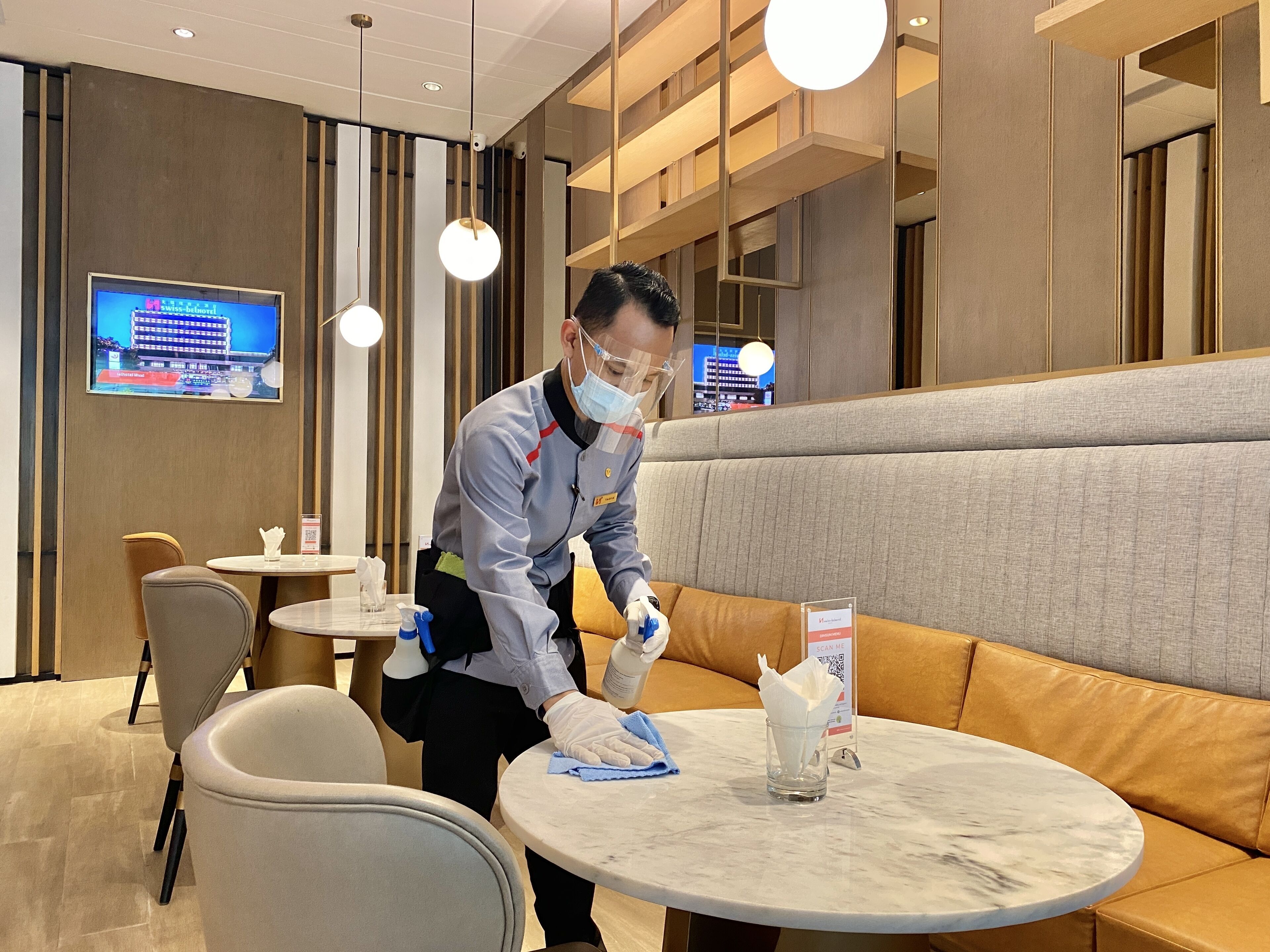 A hotel interior with a seating area, a table with a tissue dispenser, and a person cleaning a table.
