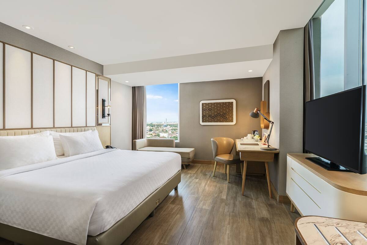 A hotel room with a large bed, a desk, a chair, a television, and a window with a city view.