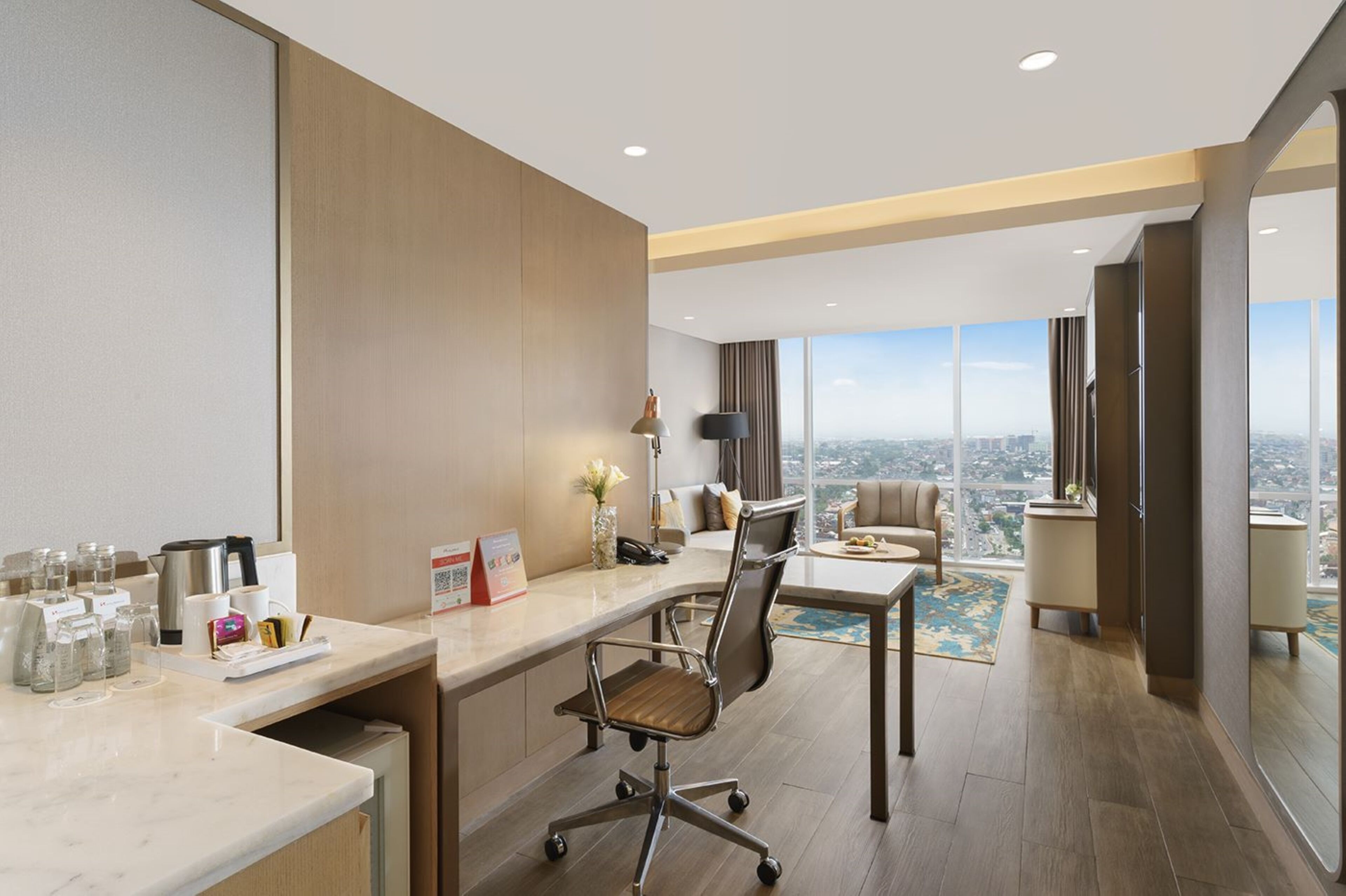 A modern hotel room with a large desk, a comfortable chair, a sofa, and a large window offering a city view.