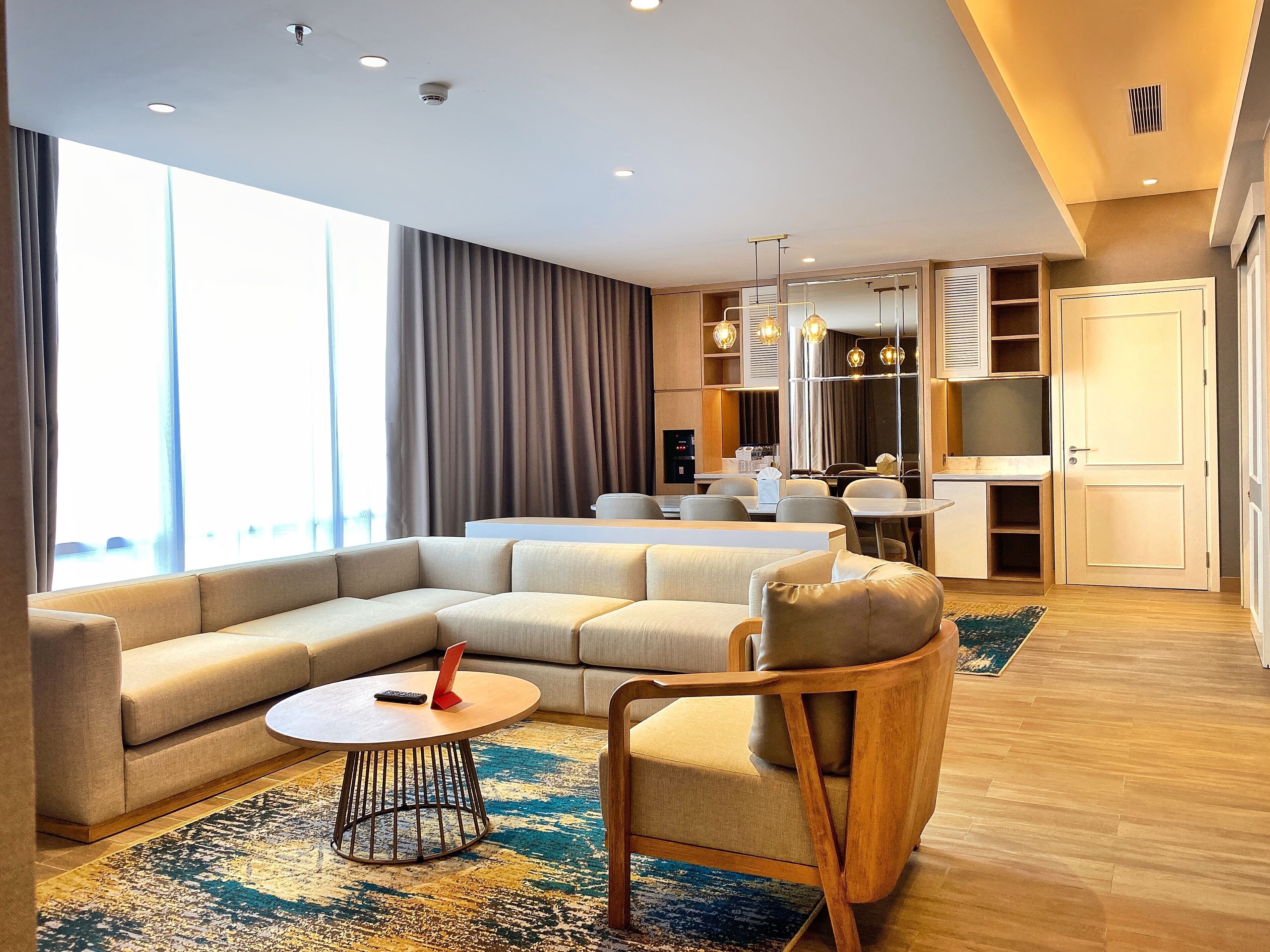 A modern hotel room with a large sectional sofa, a round coffee table, a wooden armchair, and a dining area with a small table and chairs.