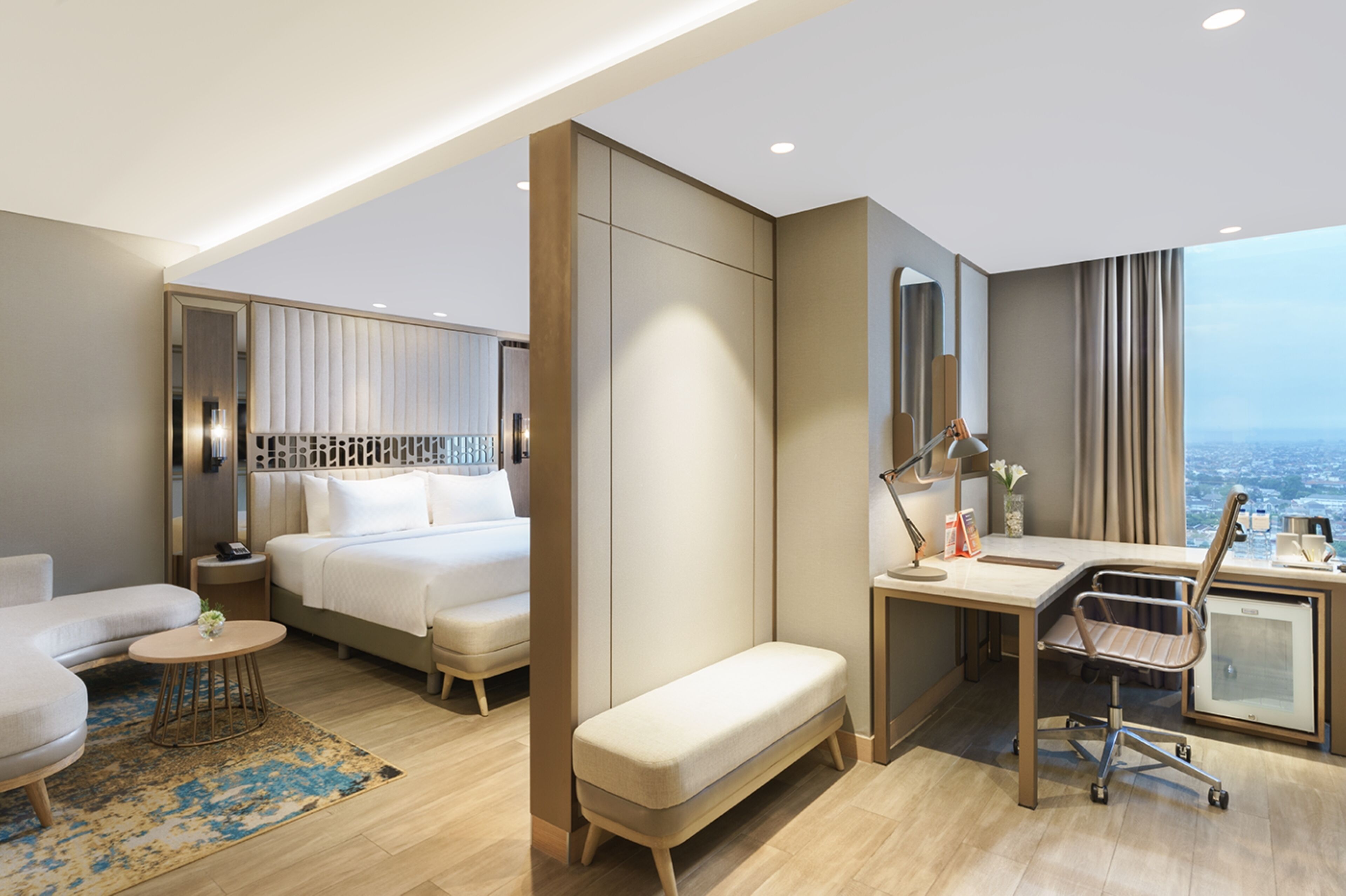 A modern hotel room with a bed, a desk, a chair, a bench, and a view of the city.