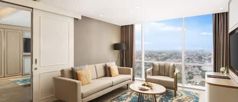 A modern living room with a sofa, armchair, and coffee table, large windows with a city view, and a flat-screen TV.