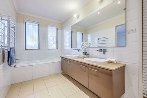 A modern bathroom with a bathtub, two sinks, a large mirror, and a window with blinds.