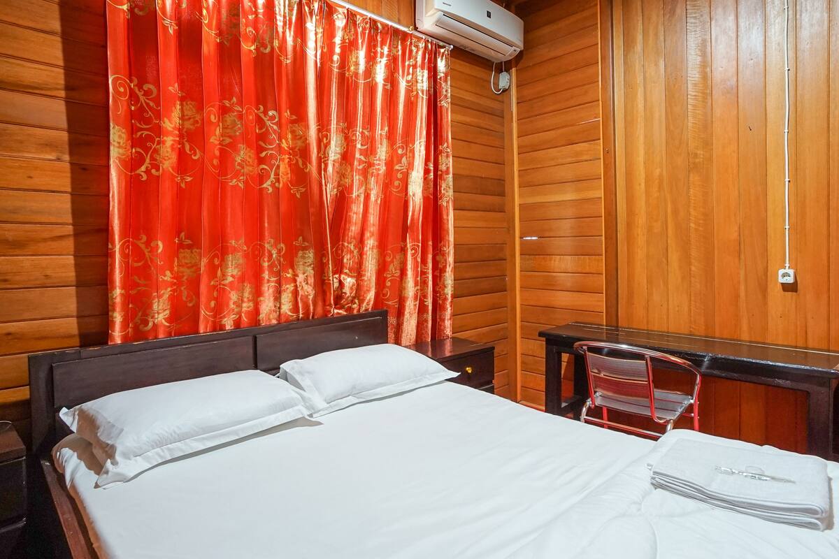 Una habitación con paneles de madera, una cama, una cortina roja, una mesita y una silla.