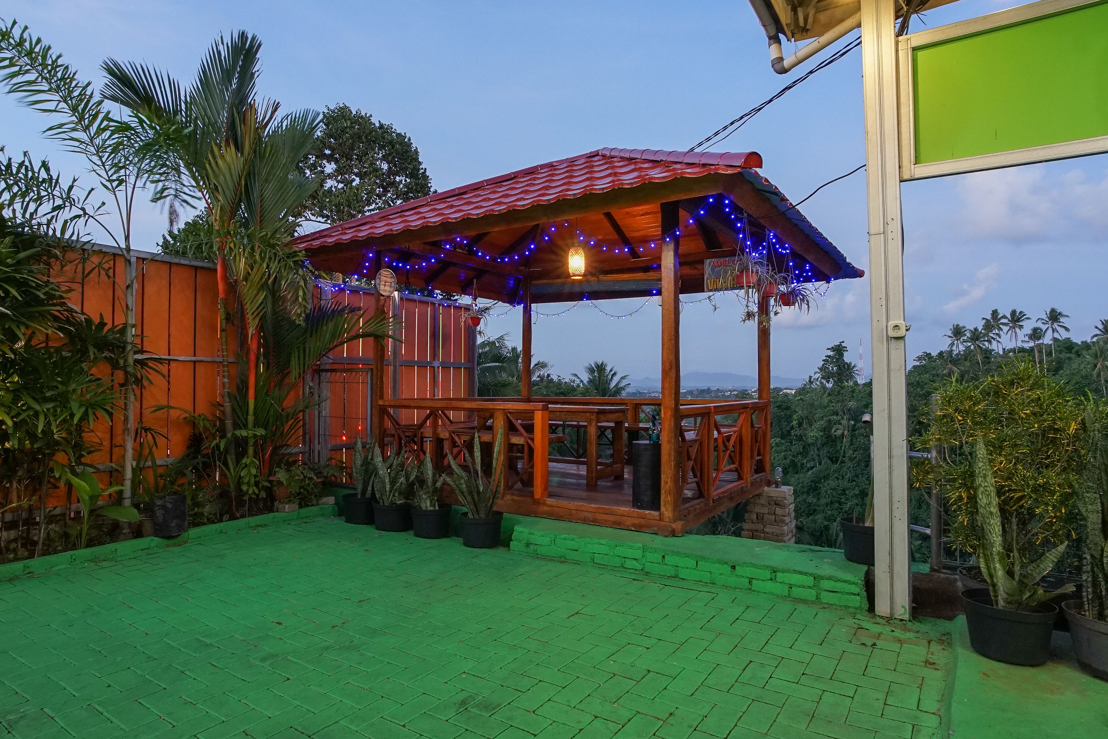 A wooden gazebo with a red roof, surrounded by potted plants and a green paved area.