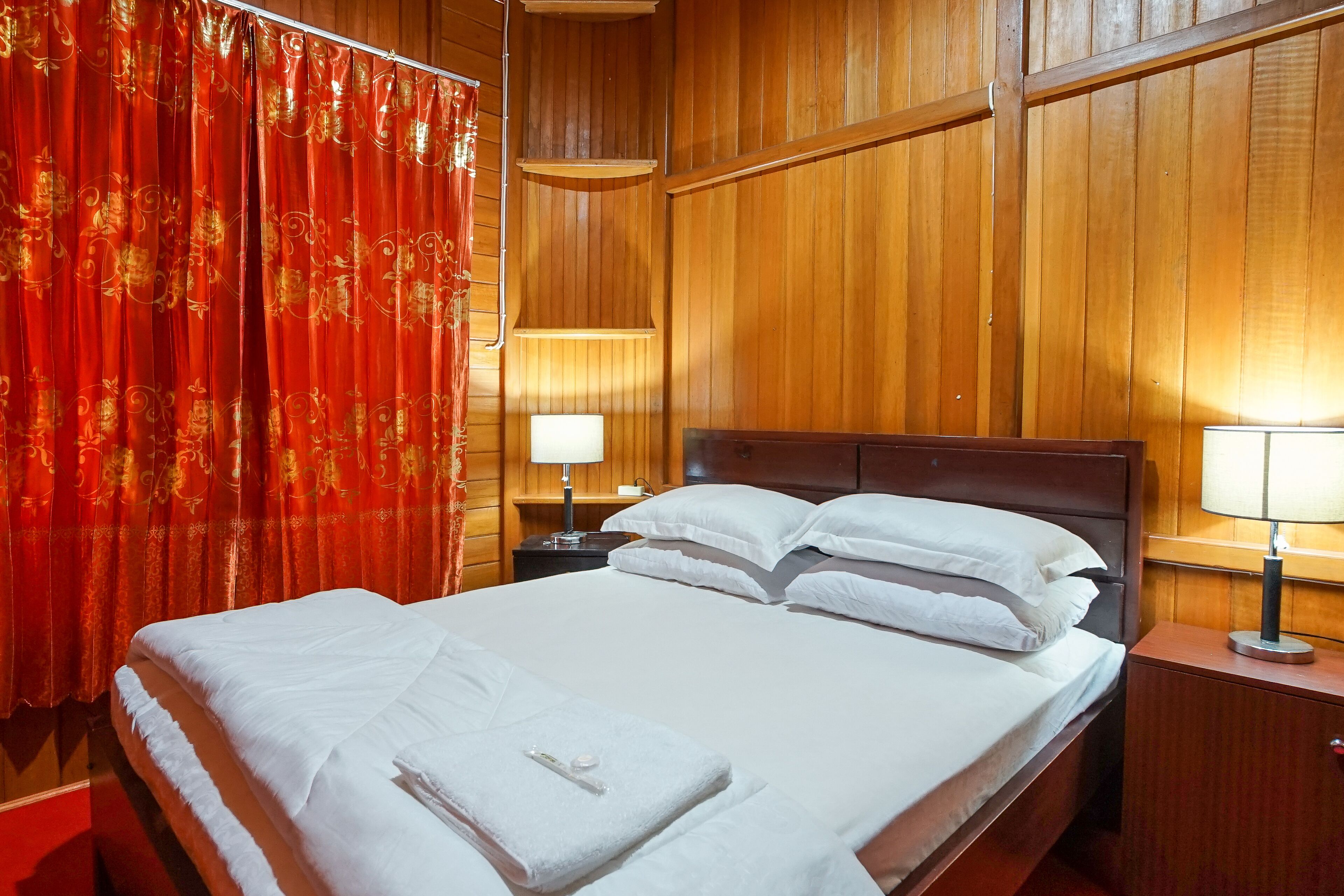 Une chambre d’hôtel avec un mur lambrissé en bois, un grand lit avec du linge de lit blanc, deux tables de chevet avec des lampes et des rideaux rouges ornés de motifs dorés.