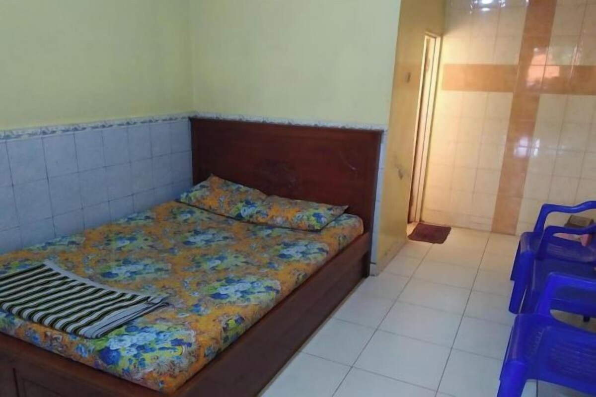 A single bed with a floral bedspread, a wooden headboard, and a blue plastic chair in a room with tiled walls and floor.