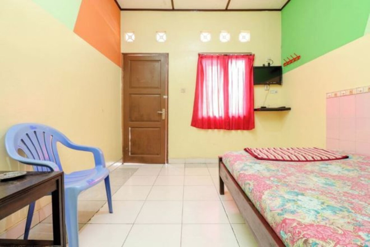 A bedroom with a bed, a chair, a desk, a door, and a wall-mounted TV.