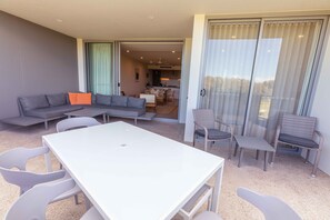 Ein modernes Wohnzimmer mit einem weißen Tisch, einem grauen Sofa und Korbstühlen. Im Hintergrund ist eine Küche zu sehen.