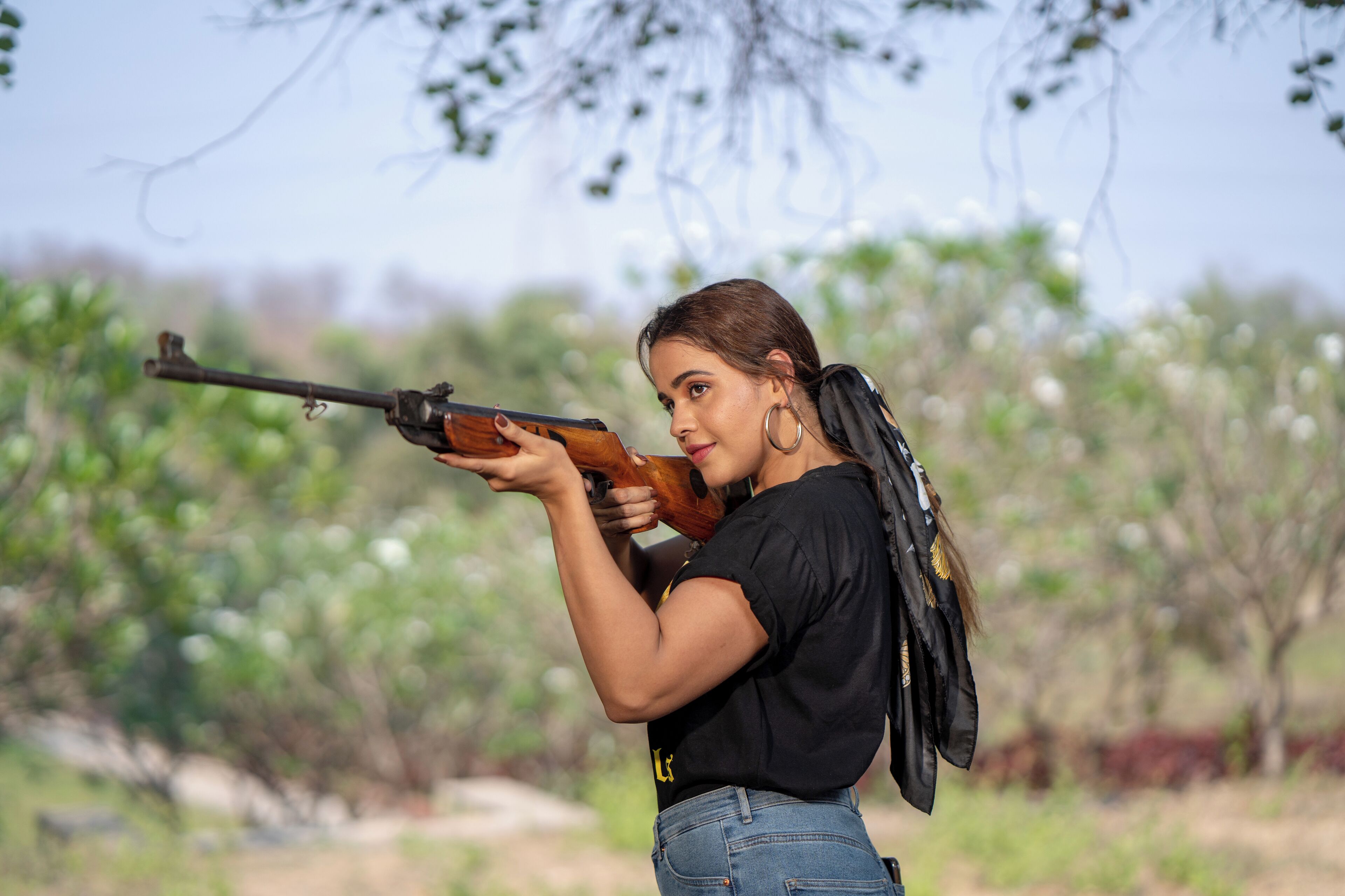 A person aiming a rifle outdoors with trees in the background.