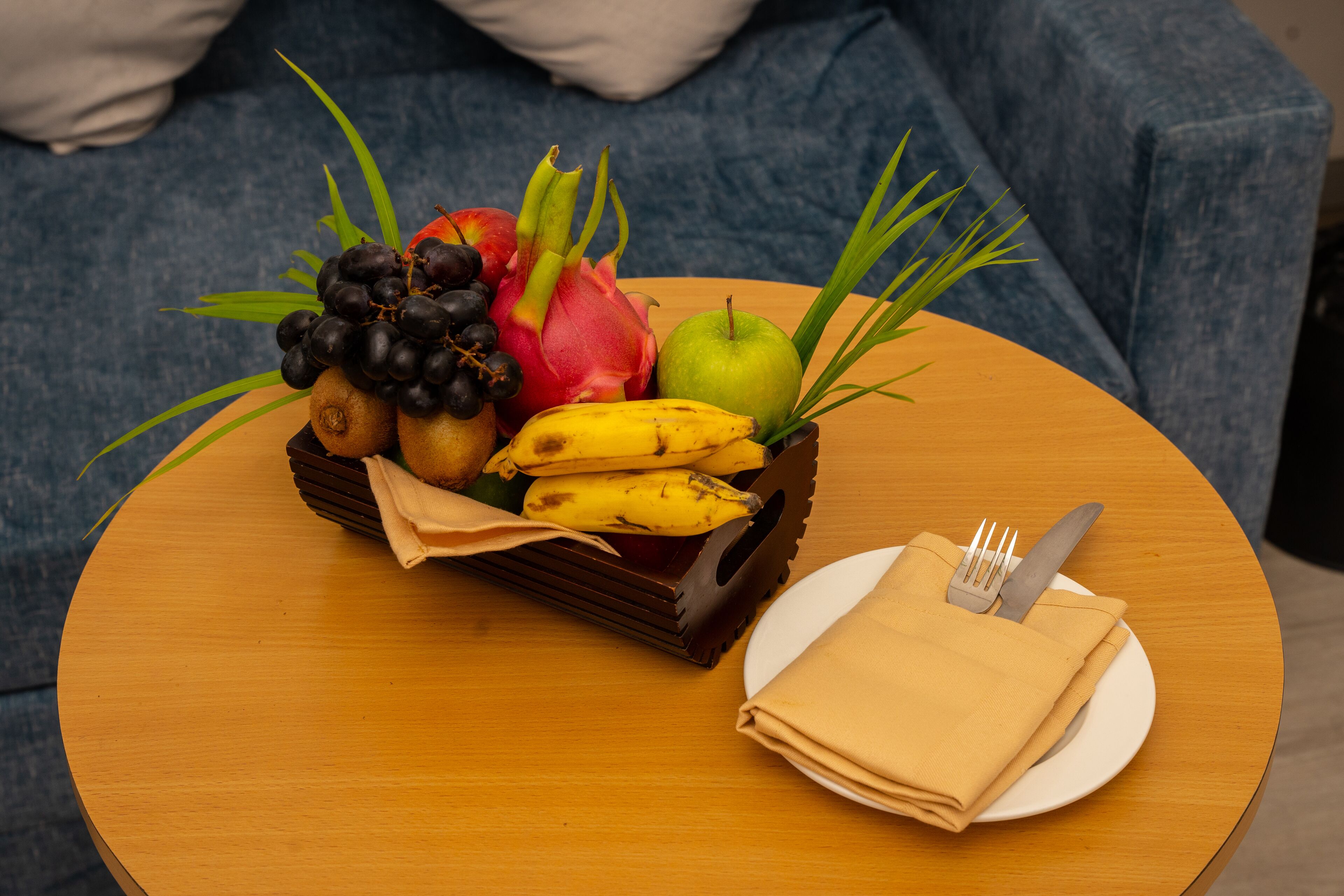 A round wooden table with a fruit basket containing grapes, bananas, a dragon fruit, and an apple, along with a folded napkin and cutlery on a white plate.