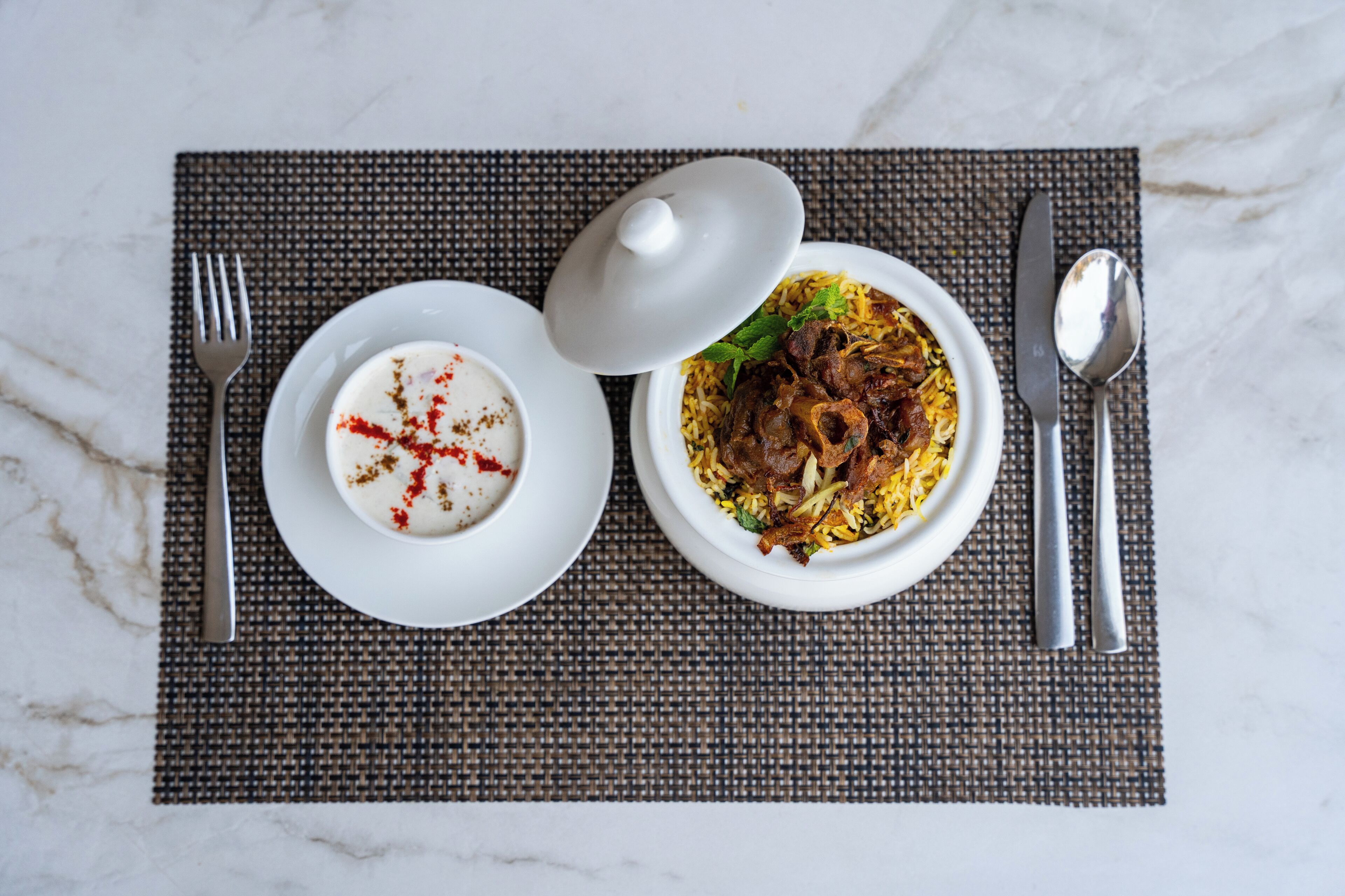 Un repas composé d’un bol de riz et de viande, d’un bol de yaourt agrémenté d’une épice rouge, et d’une assiette blanche avec une cuillère et une fourchette, le tout posé sur un set de table texturé.