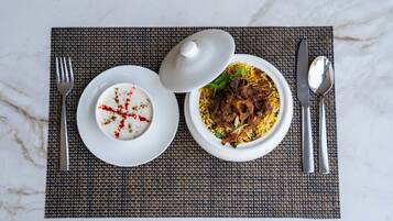 Un repas composé d’un bol de riz et de viande, d’un bol de yaourt agrémenté d’une épice rouge, et d’une assiette blanche avec une cuillère et une fourchette, le tout posé sur un set de table texturé.