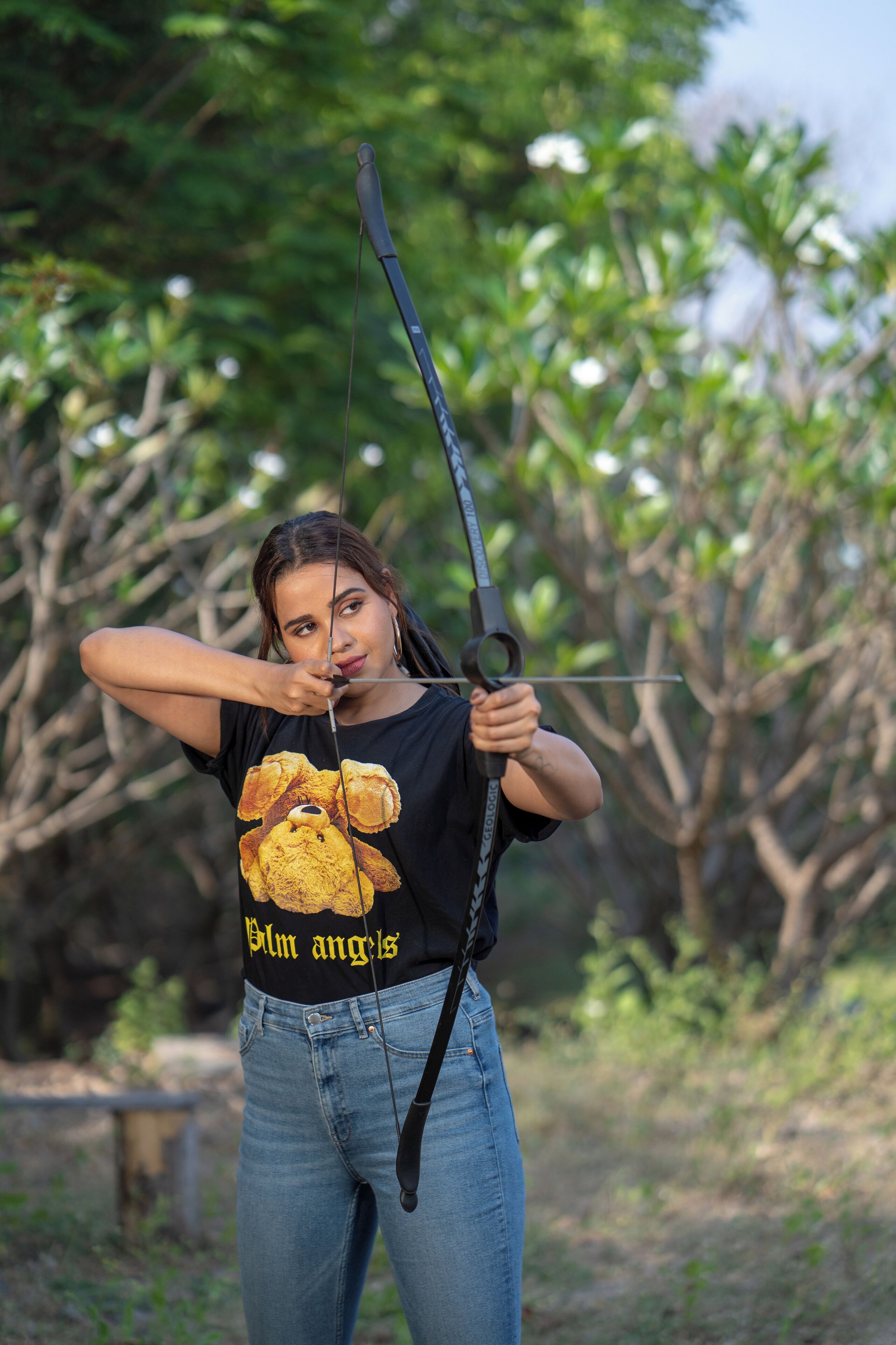 Una persona sosteniendo un arco y una flecha al aire libre, vistiendo una camiseta negra con un diseño y pantalones vaqueros azules.