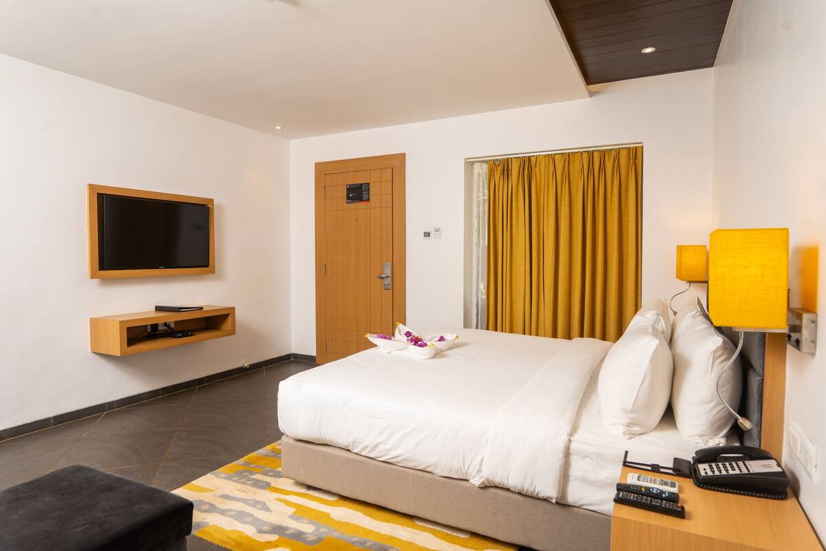 Una habitación de hotel con cama, televisor montado en la pared, mesita de noche con lámpara y puerta con panel de madera.