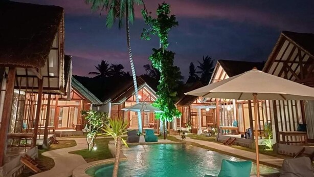 A resort with a swimming pool, thatched-roof buildings, and a palm tree. - LMBK Surf Camp - Hostel (Kuta)