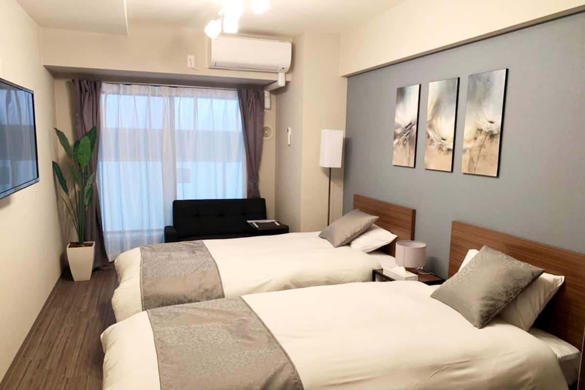 A hotel room with two beds, a large window with curtains, a potted plant, and artwork on the wall.