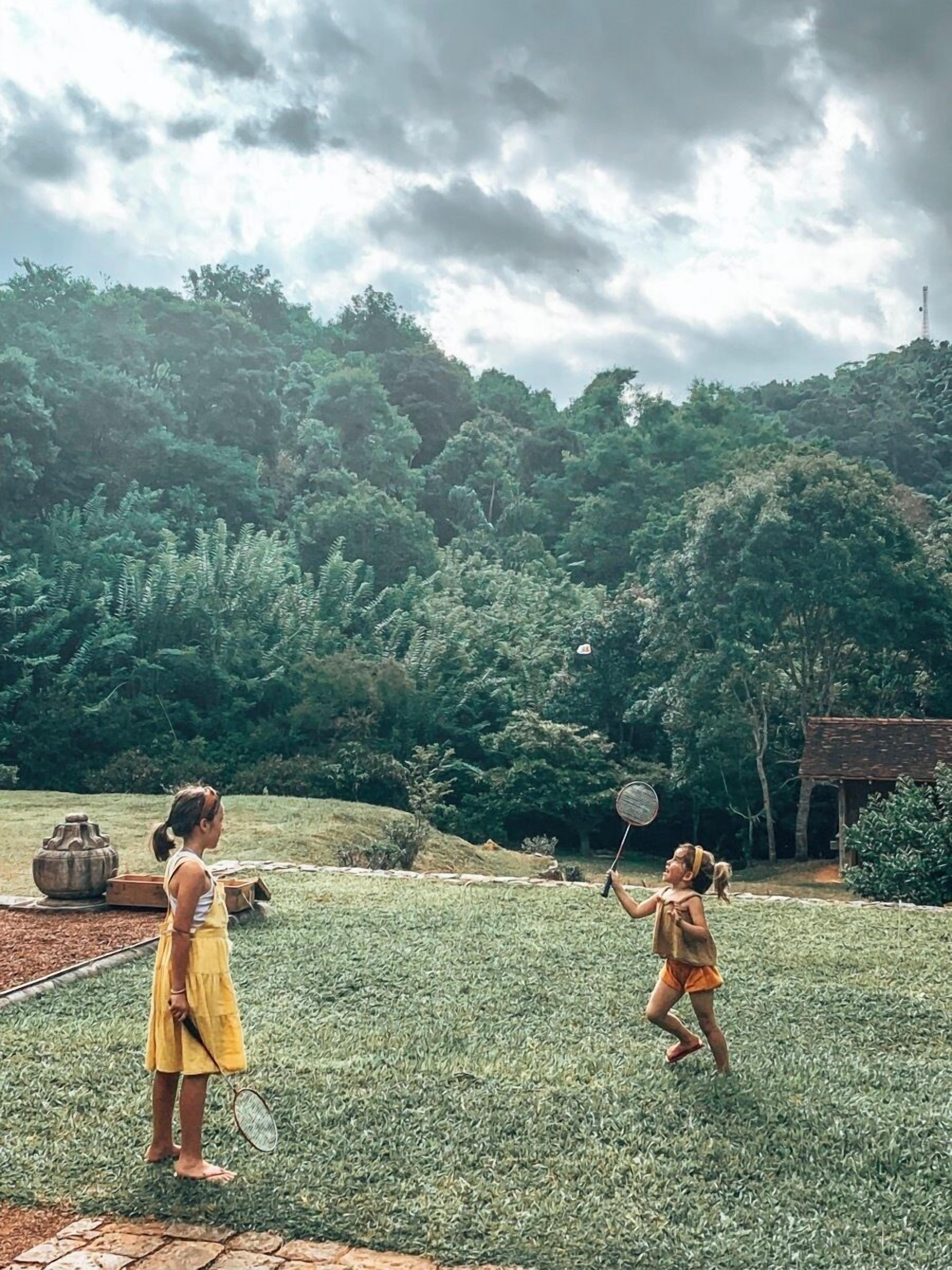 Two individuals playing badminton in a grassy outdoor area with a backdrop of dense trees and a cloudy sky.