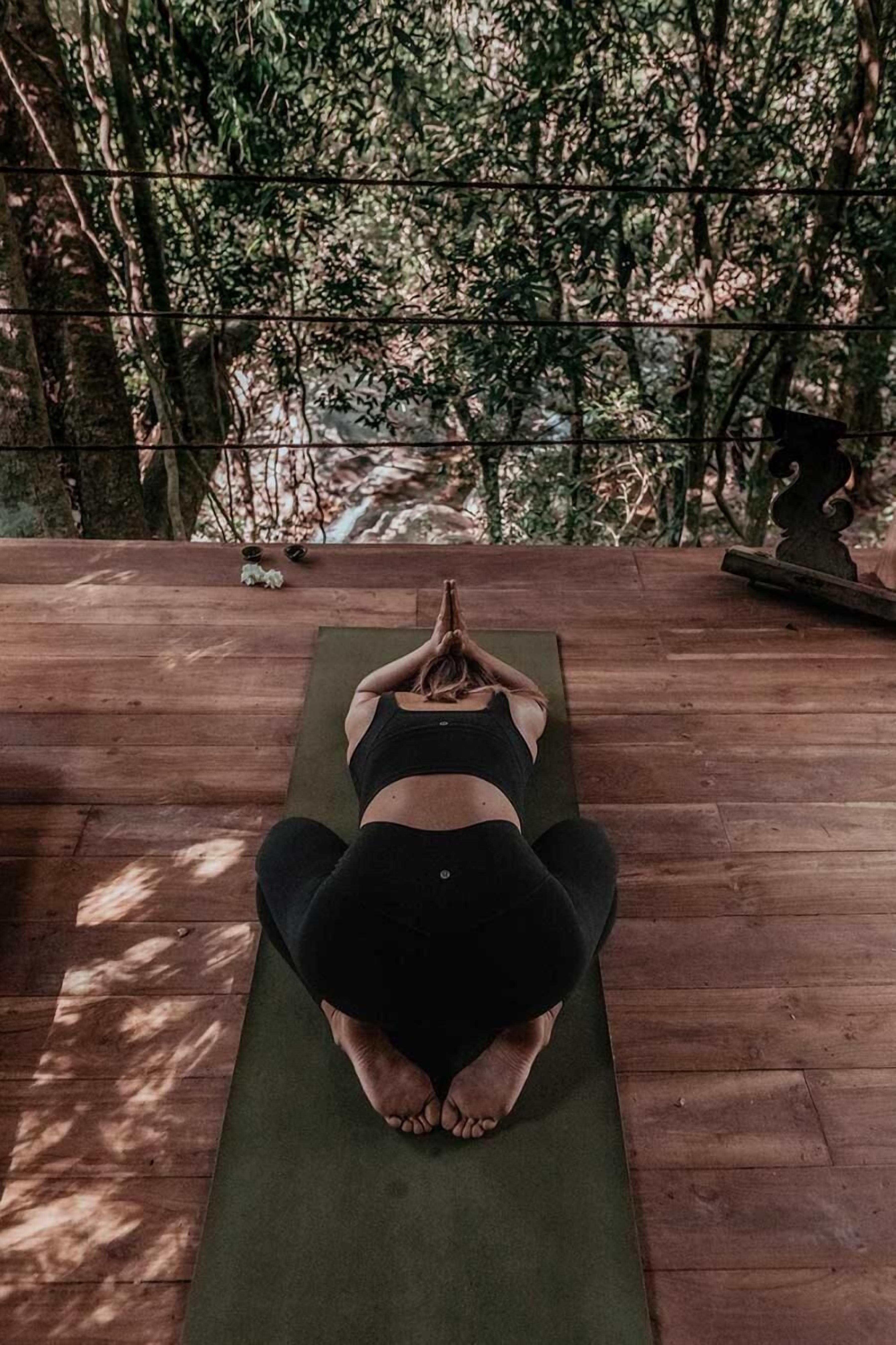 A person performing a yoga pose on a mat in a room with wooden flooring and a view of a forested area.