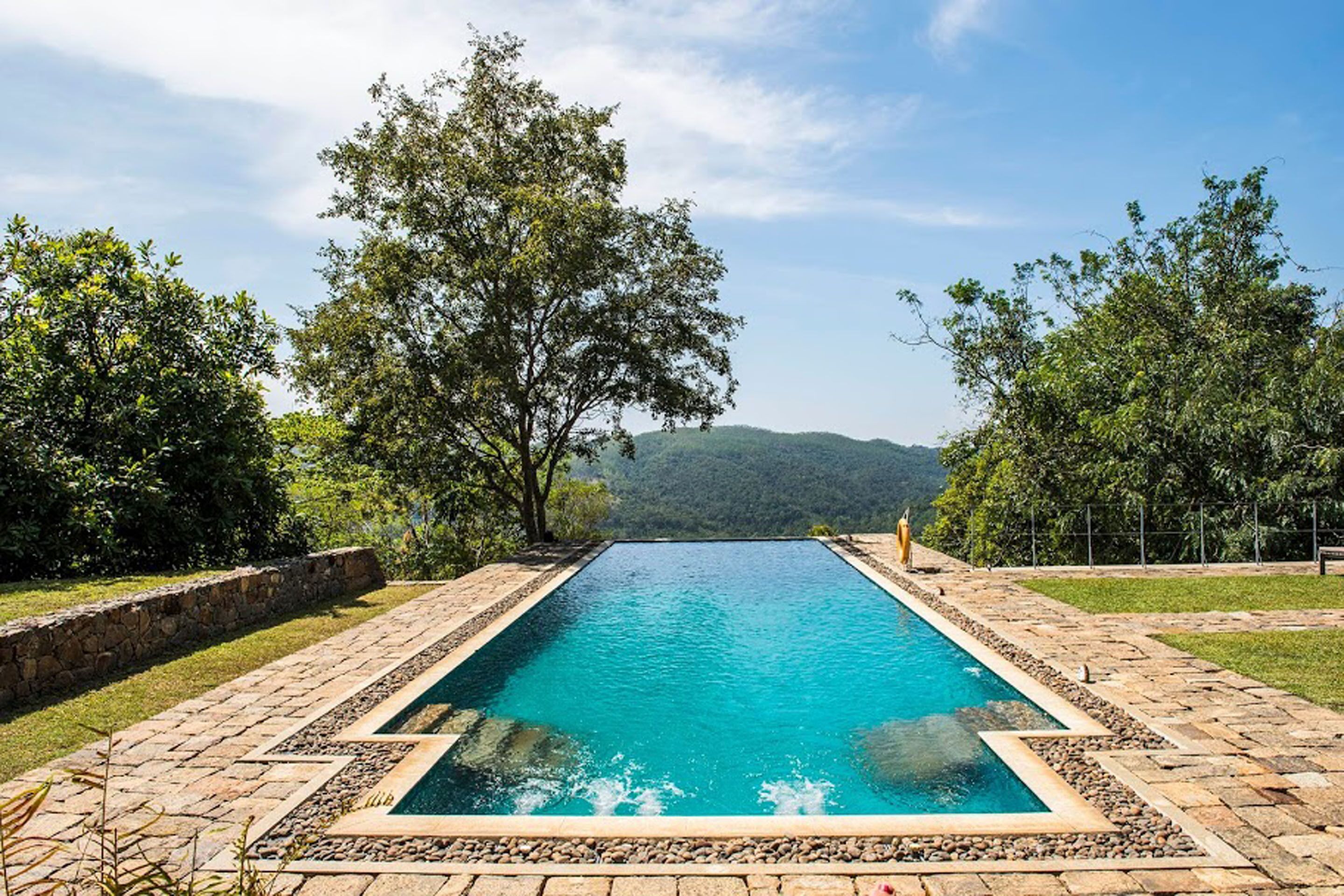Uma piscina retangular com borda de pedra, rodeada por árvores e um muro de pedra, com vista para colinas ao fundo.