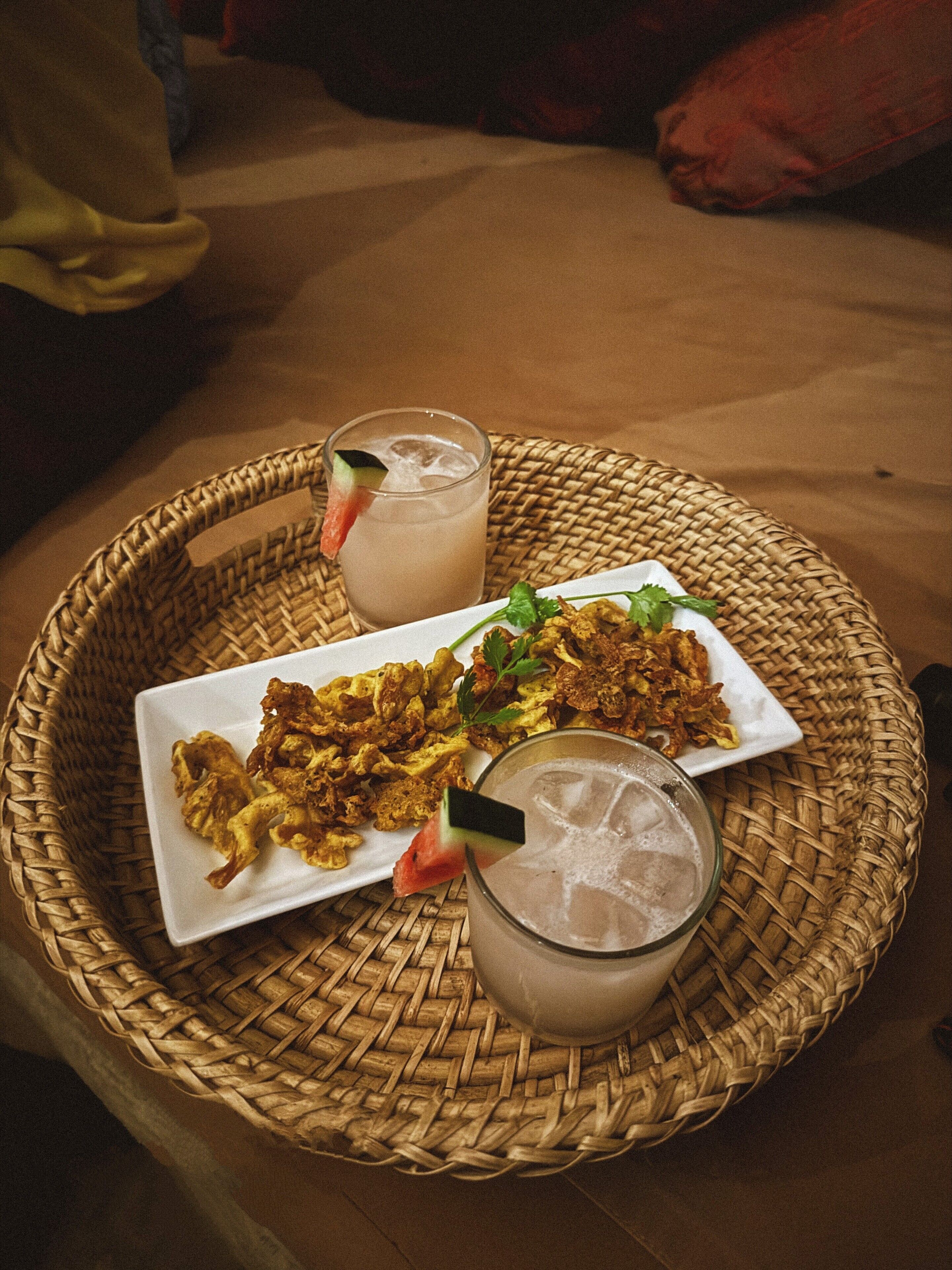 A woven tray with a plate of fried food and two glasses of a beverage with ice and a slice of fruit.