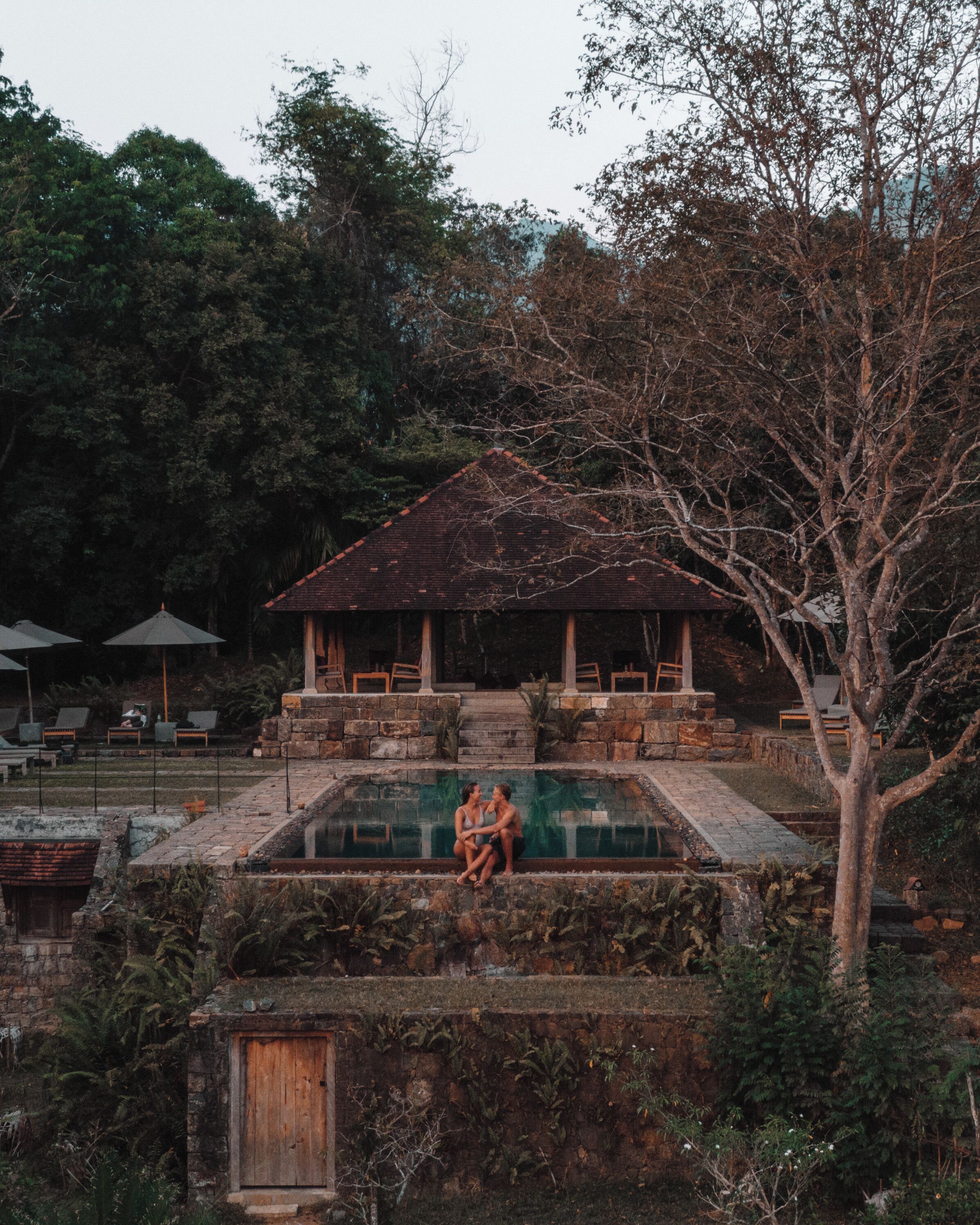 A stone pool with two people sitting at the edge, surrounded by lush greenery and a pavilion with a tiled roof.