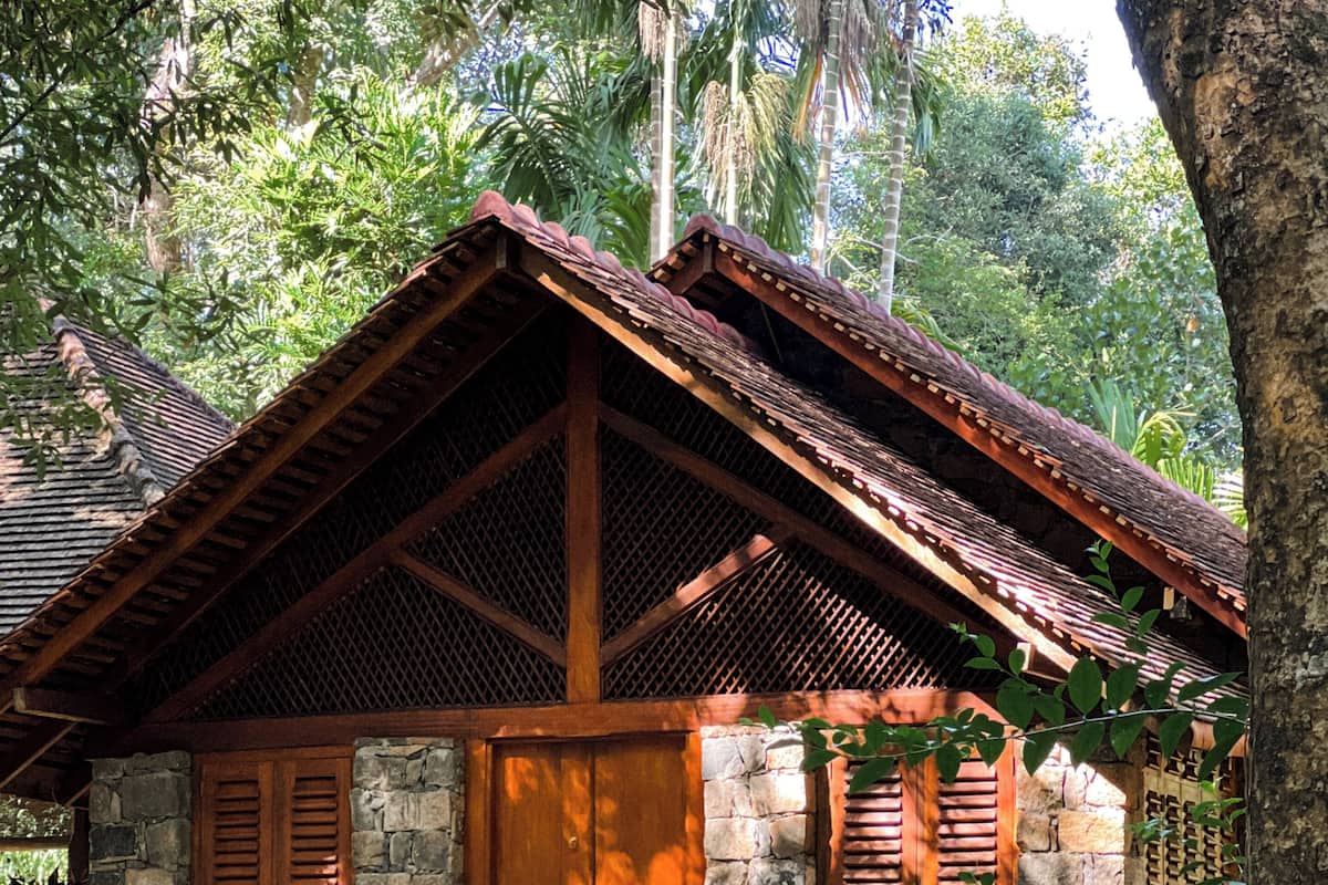 A rustic stone building with a wooden door and shutters, surrounded by lush greenery.