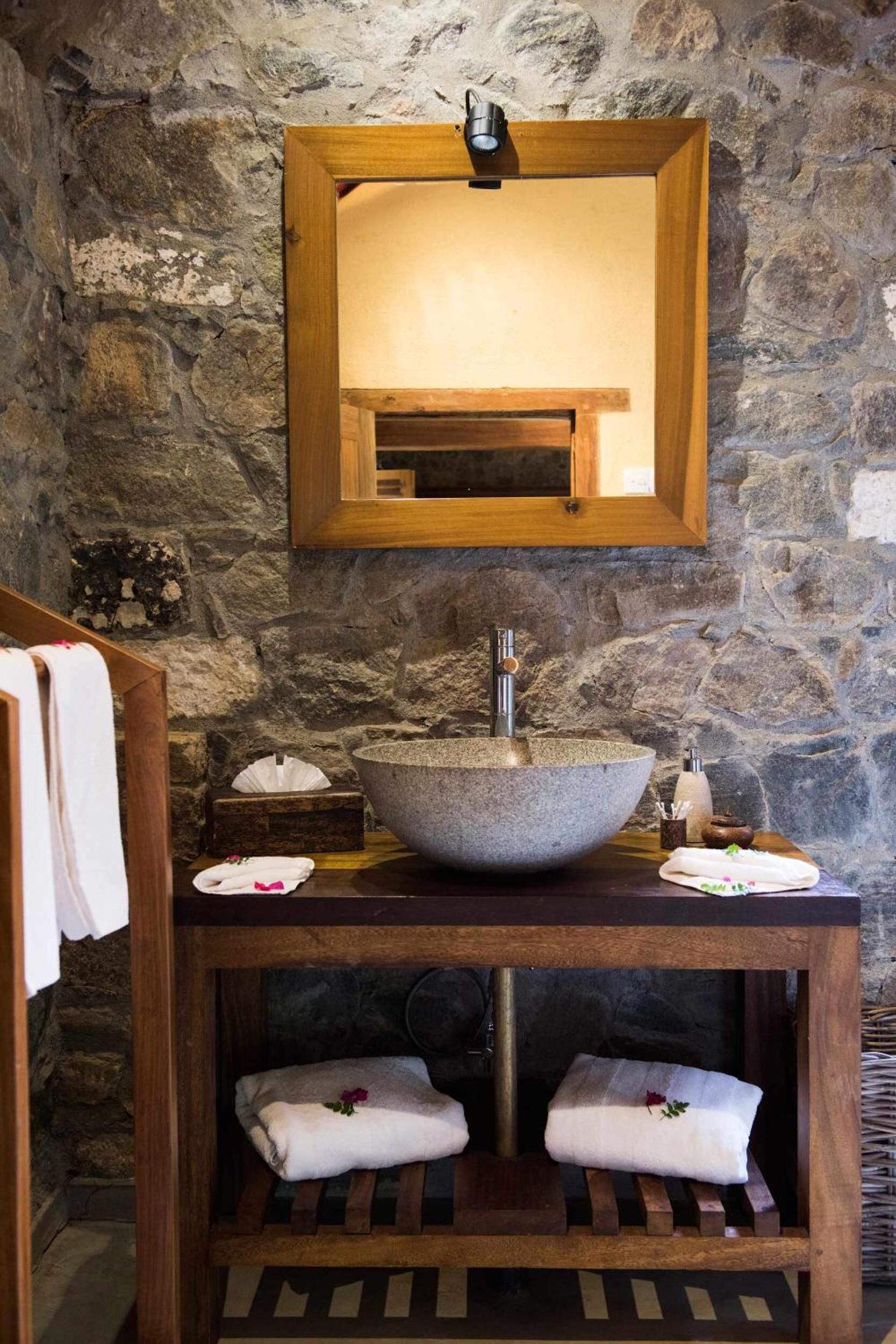 Bagno con parete in pietra, mobile lavabo in legno, un ampio lavabo a ciotola e uno specchio sopra.