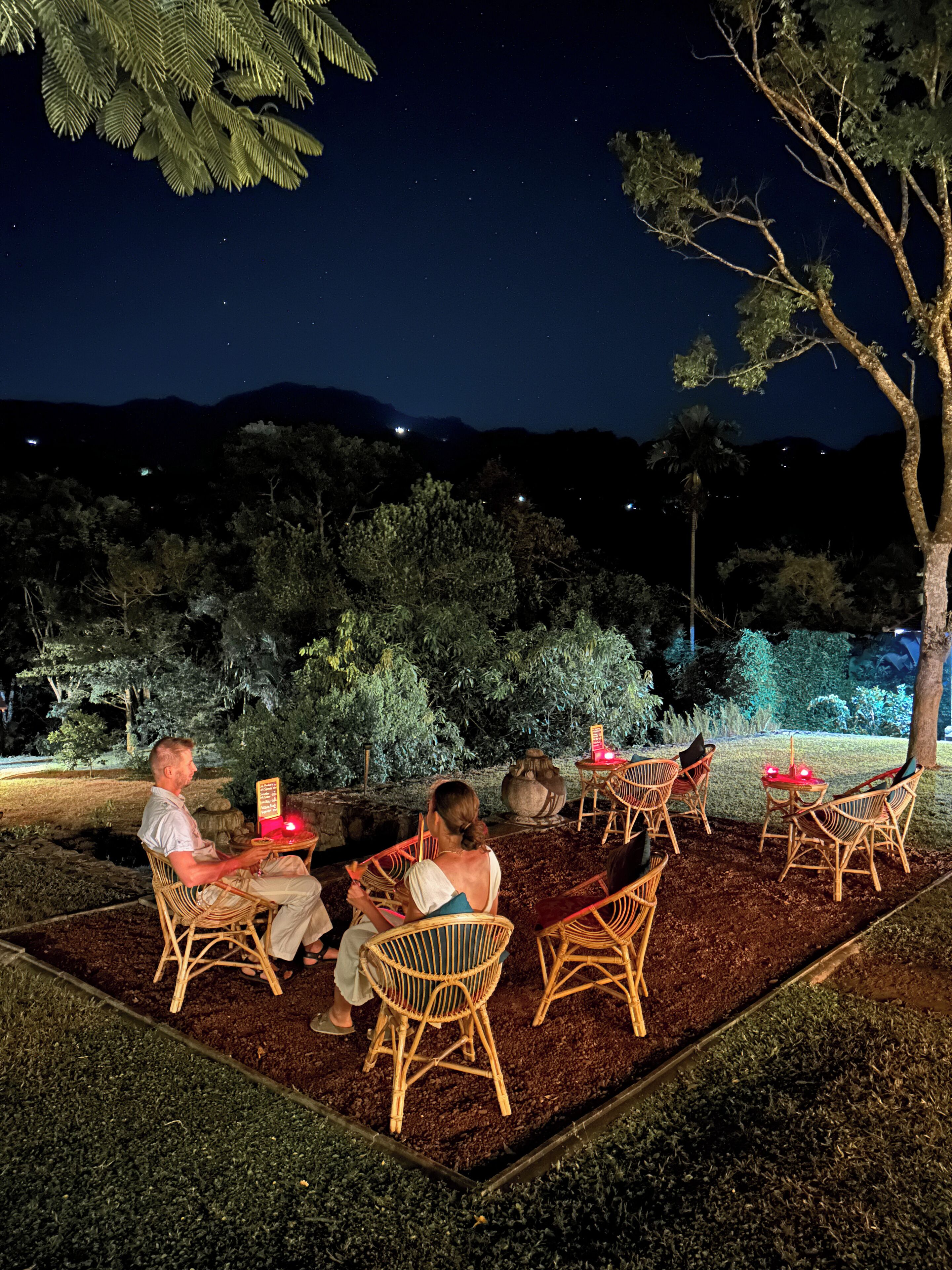 An outdoor seating area with wicker chairs and tables, surrounded by trees and a view of the night sky.
