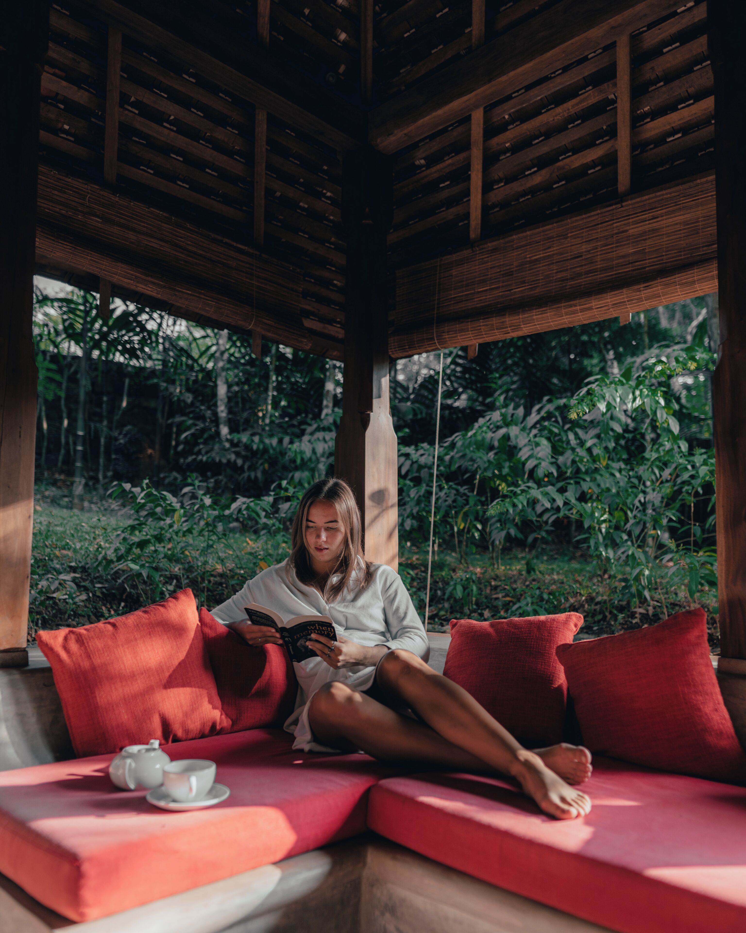 Uma pessoa sentada em um banco vermelho acolchoado, lendo um livro em uma área externa coberta, com vigas de madeira e vegetação exuberante ao fundo.