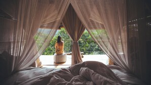 Ein Zimmer mit einem Bett, eine Person sitzt am Fenster und blickt auf viel Grün.