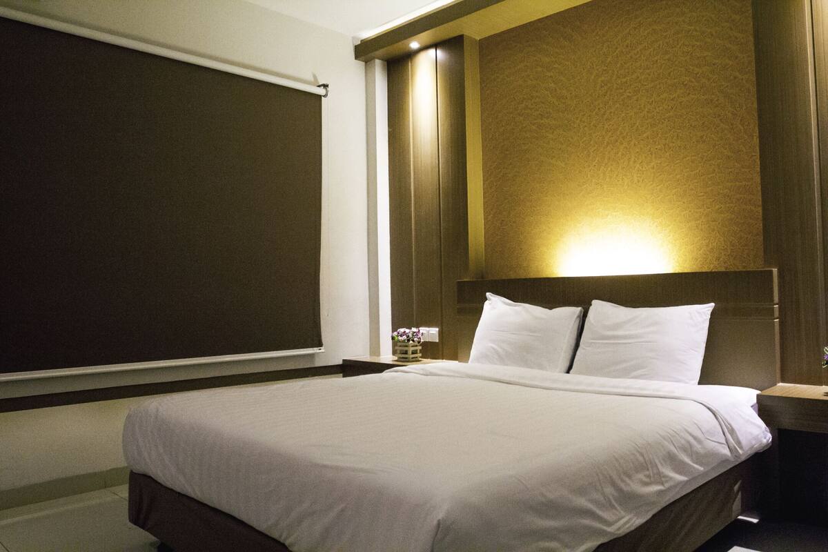 Una habitación de hotel con una cama grande, cabecera con luz y una ventana con persiana.