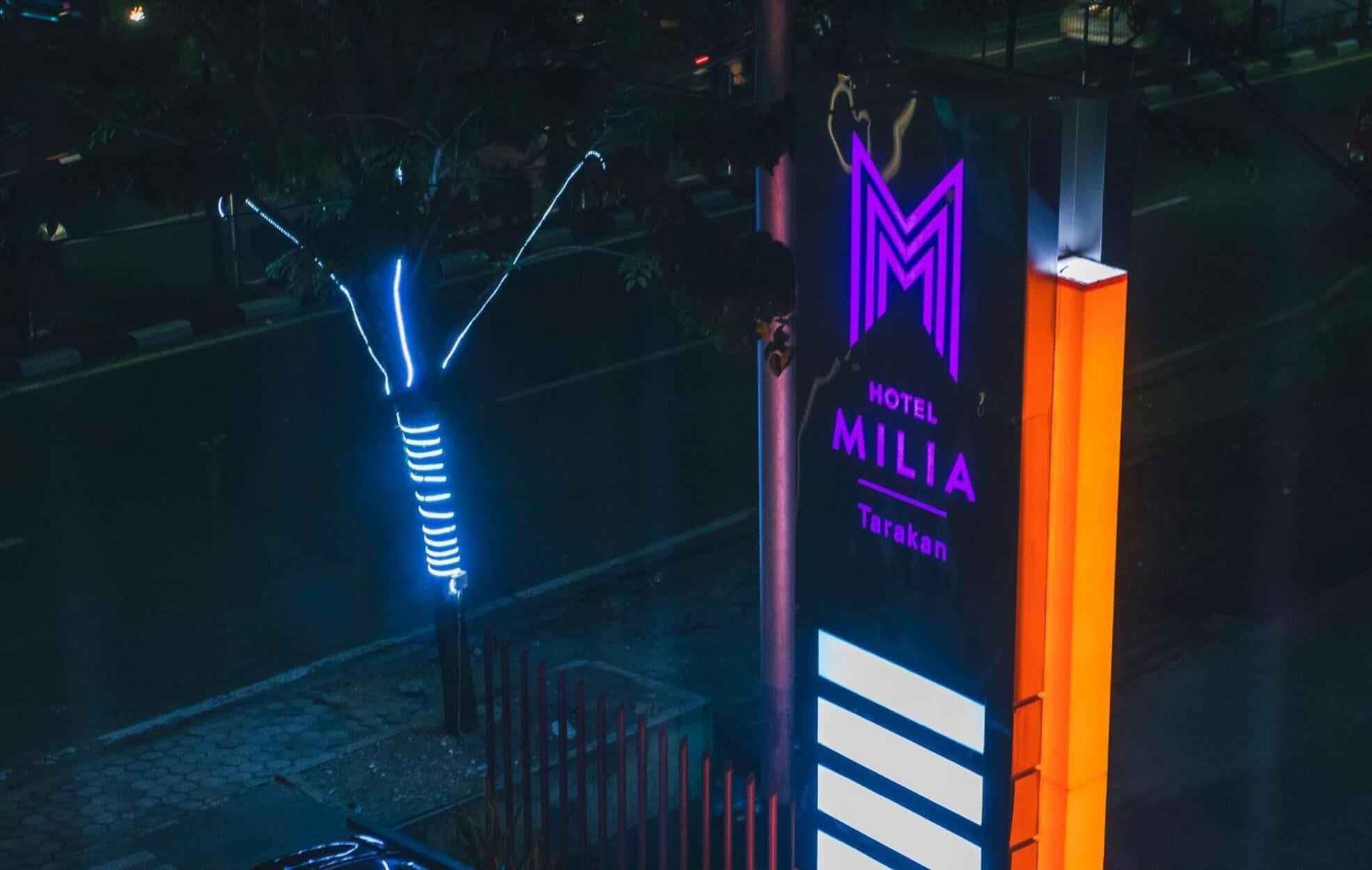 Letrero de neón del Hotel Milia en Tarakan, que presenta un logotipo estilizado con la letra 'M' y un cartel vertical con el nombre y la ubicación del hotel.