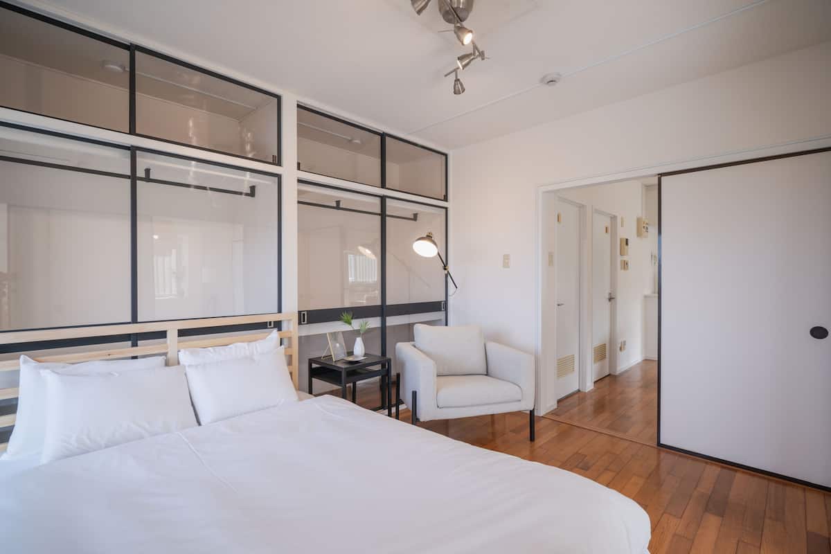 A modern hotel room with a large bed, a small table with a lamp, a white armchair, and a large window.