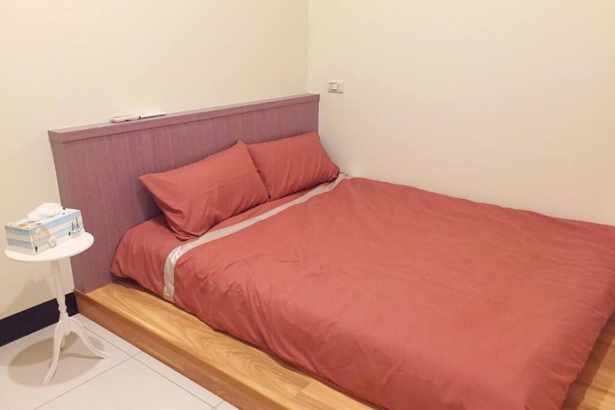 A single bed with a wooden headboard and a red bedspread. There is a small white table with a box on it to the left of the bed.