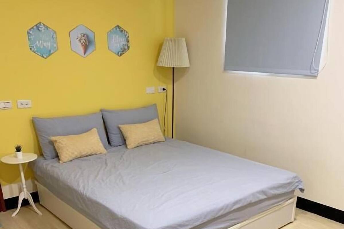 A neatly made bed in a room with a yellow wall, a small bedside table, and a lamp.