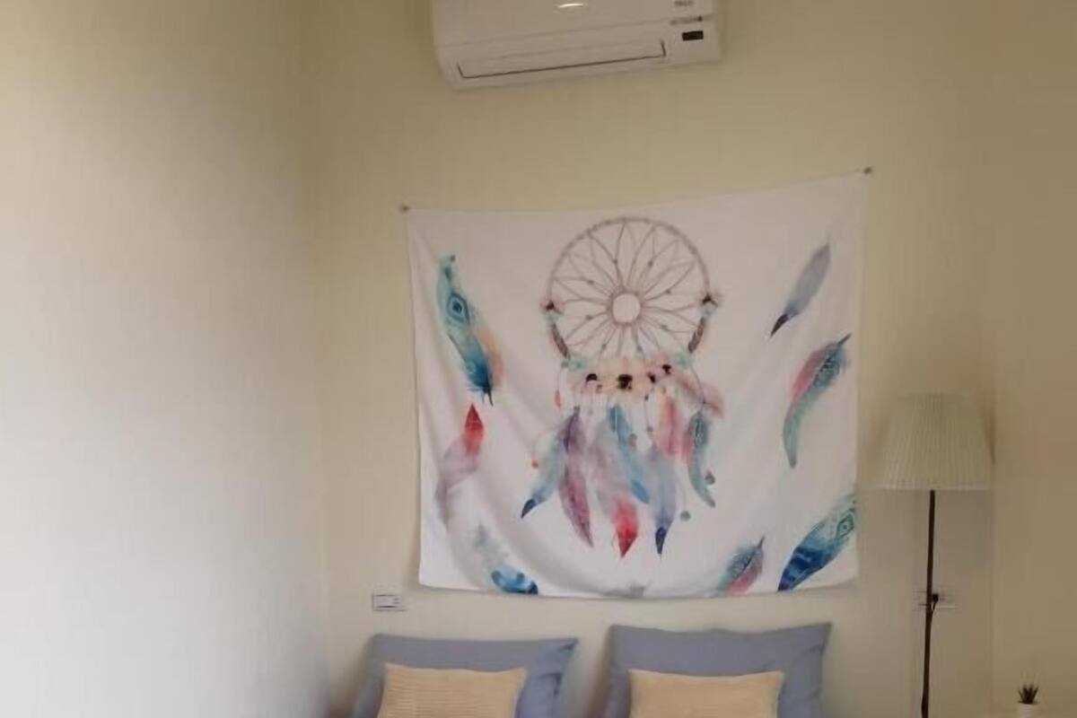 A bedroom with a bed, a wall-mounted air conditioner, and a tapestry with a dreamcatcher design.