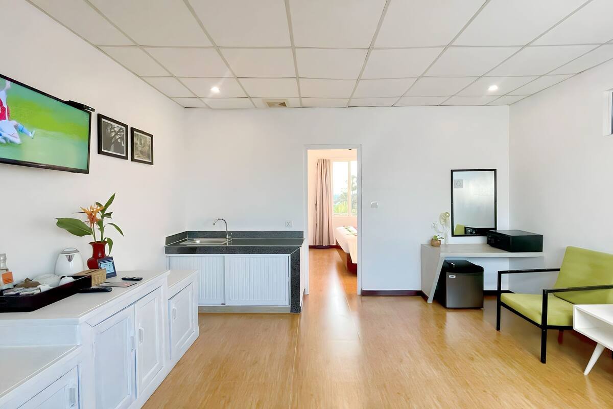 Habitación de hotel moderna con cocina, una mesa de comedor, una silla verde y un televisor mostrando un partido deportivo.