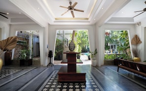 Um hall amplo com uma estátua central de Buda, um ventilador de teto e vista para um jardim exuberante através de grandes janelas.