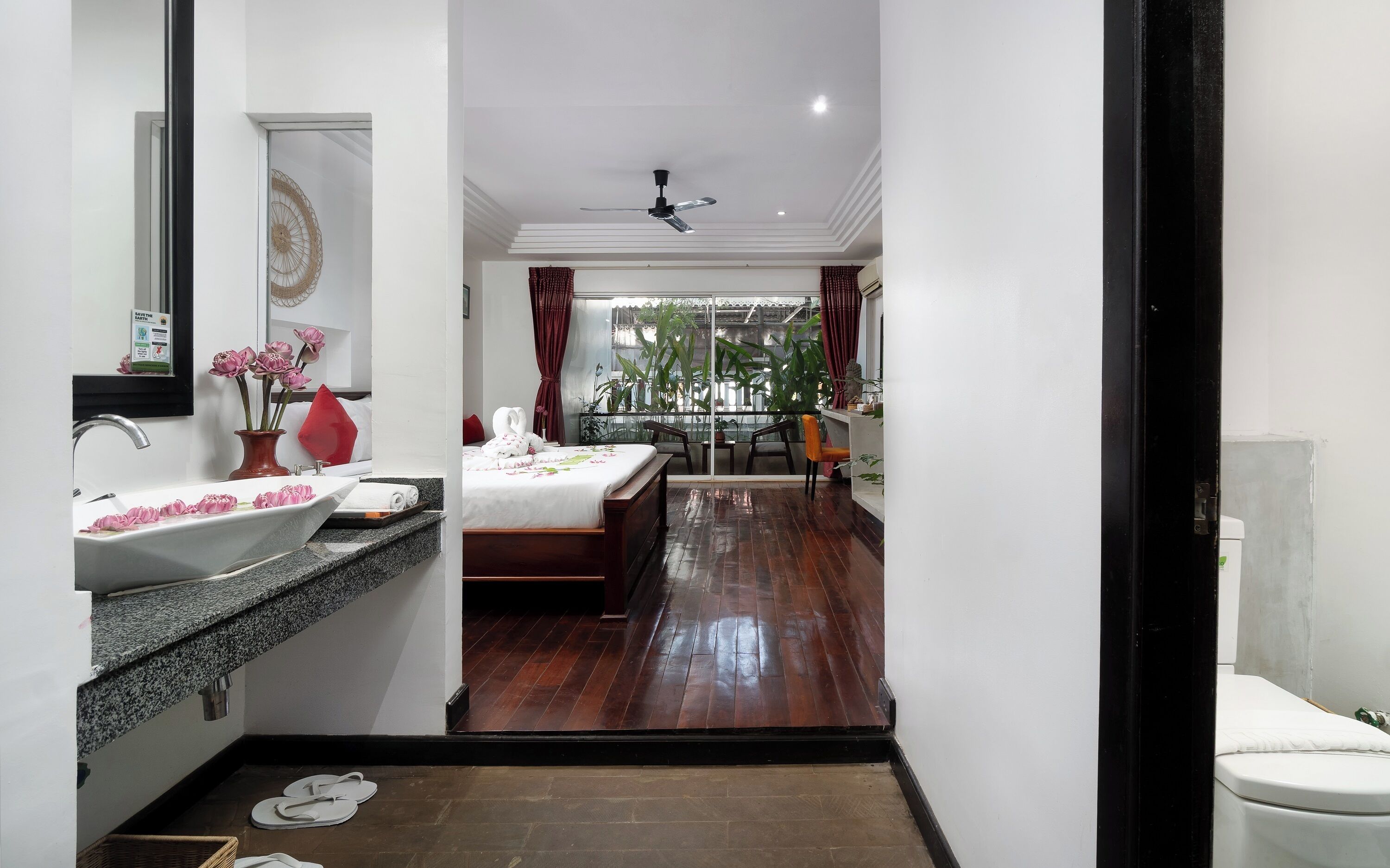 A hotel room with a bathroom, a balcony with plants, a bed, and a dining area.